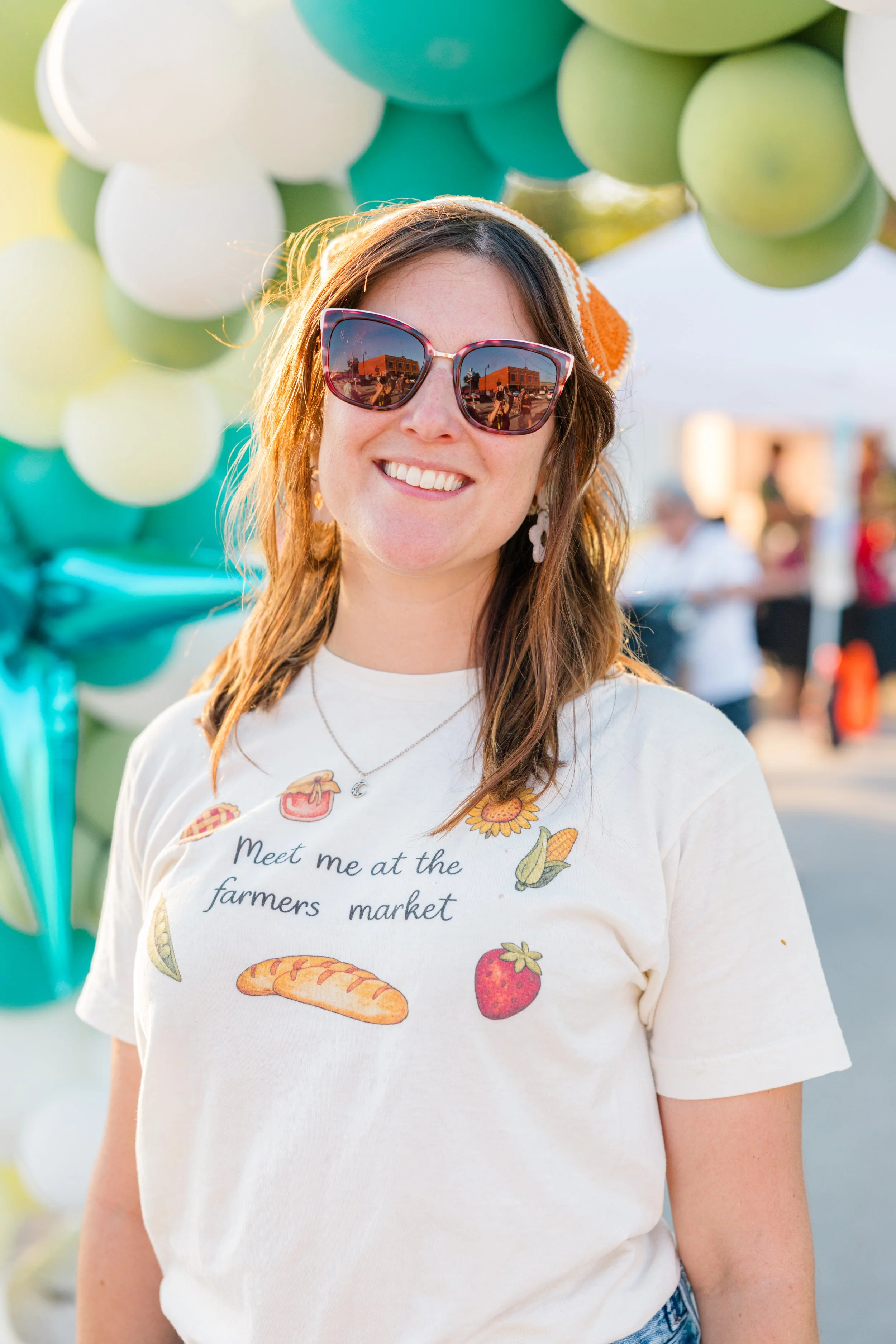 Two people sitting at a table at an outdoor farmers market, smiling, with tote bags reading 'Valley View Farmers Market' in front of them, in a park setting with a gazebo in the background.