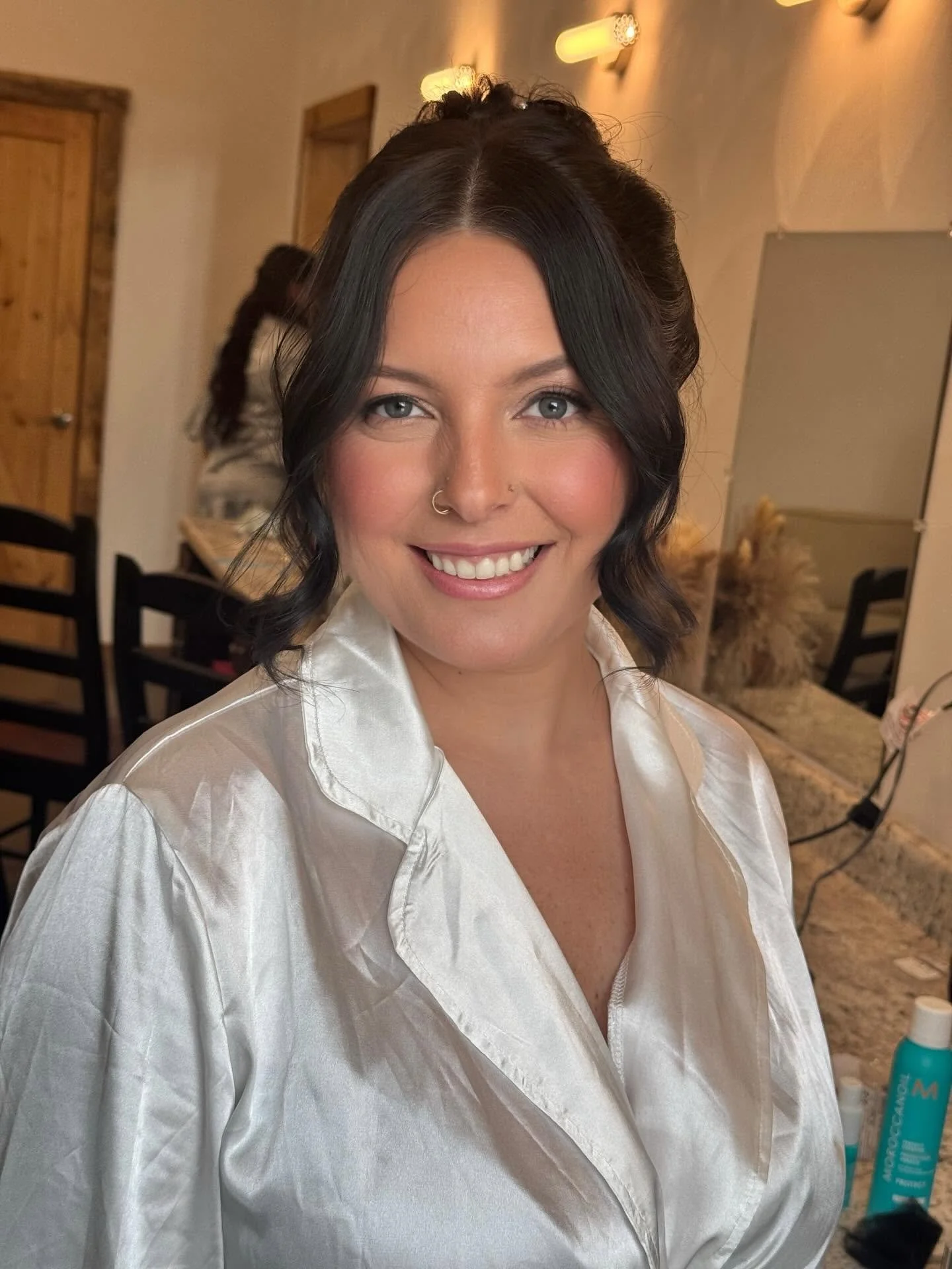 A woman with dark hair styled in loose waves, wearing a white satin robe, smiling at the camera in a warmly lit room.