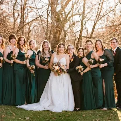 Group of women in formal dresses, including a bride in a white wedding gown holding a bouquet, standing outdoors in front of trees with autumn foliage.