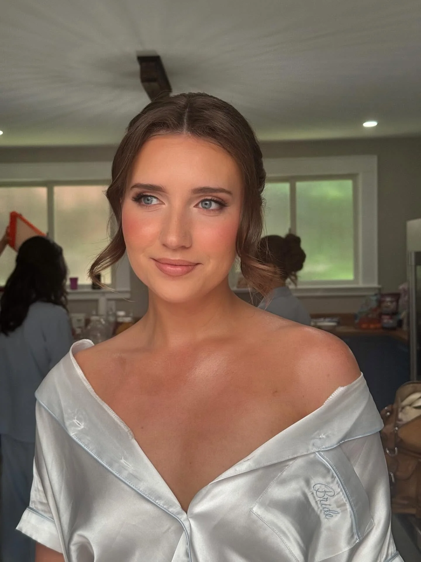 A woman with blue eyes and brown hair styled in loose curls wearing a satin off-the-shoulder white dress, standing in a room with people and large windows in the background.