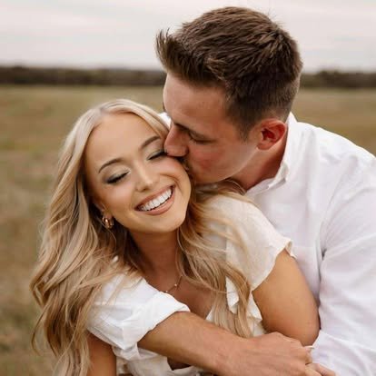 A smiling woman with blonde hair hugging a man who is kissing her on the cheek outdoors in a field.
