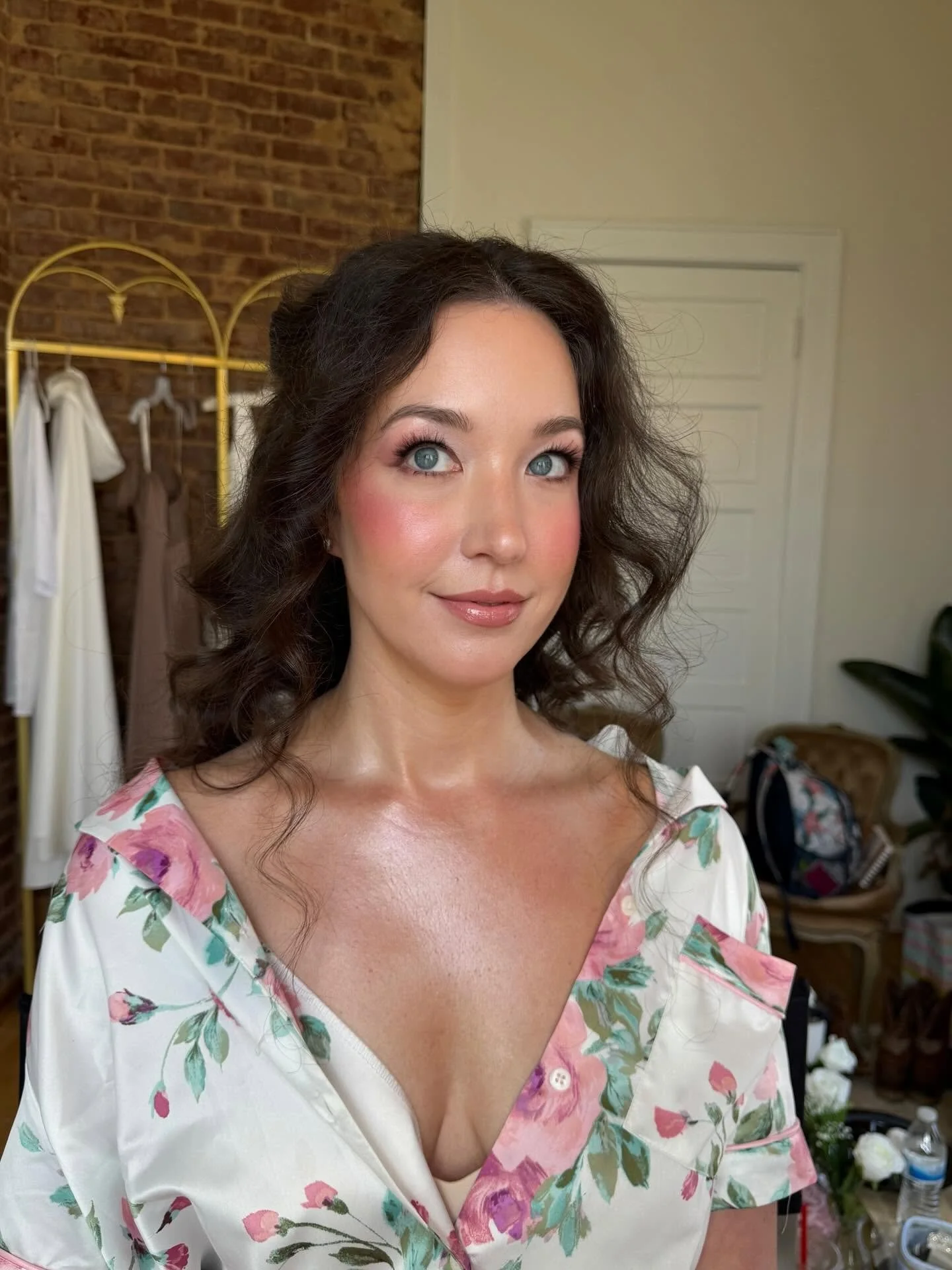 A woman with dark curly hair and blue eyes smiling while wearing a floral dress with pink, green, and white colors. She is indoors with a brick wall and a white door behind her, and some clothing and household items in the background.