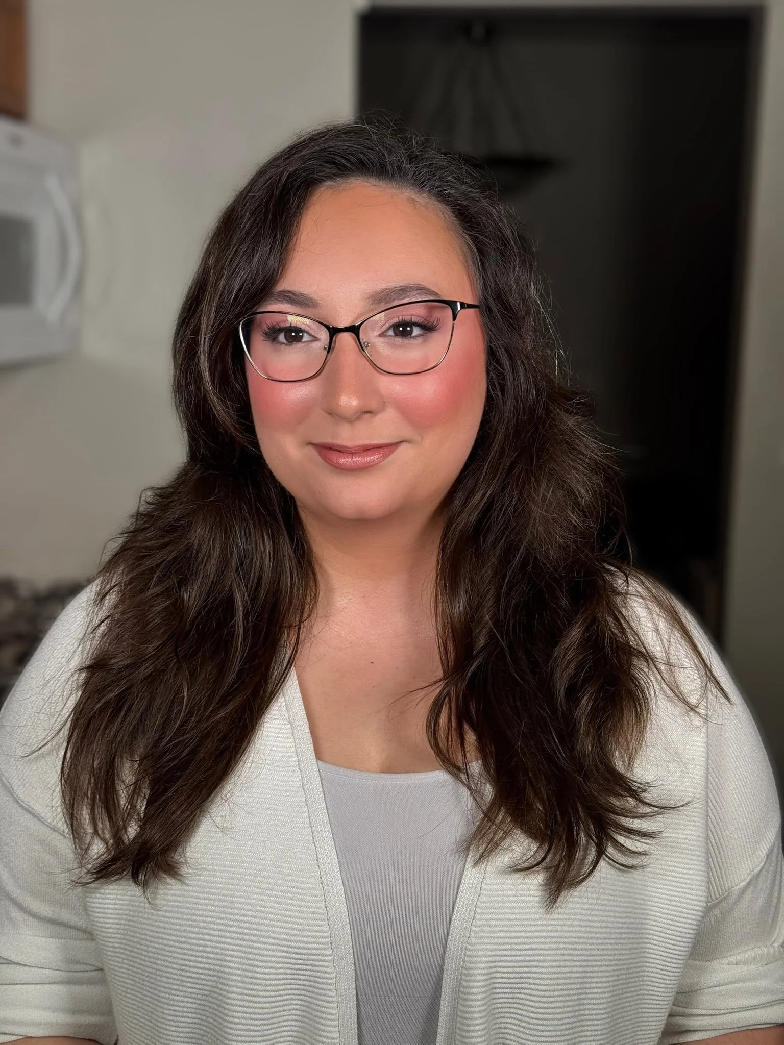 A woman with long dark hair, glasses, and a warm smile wearing a light-colored cardigan and a white top, standing indoors.