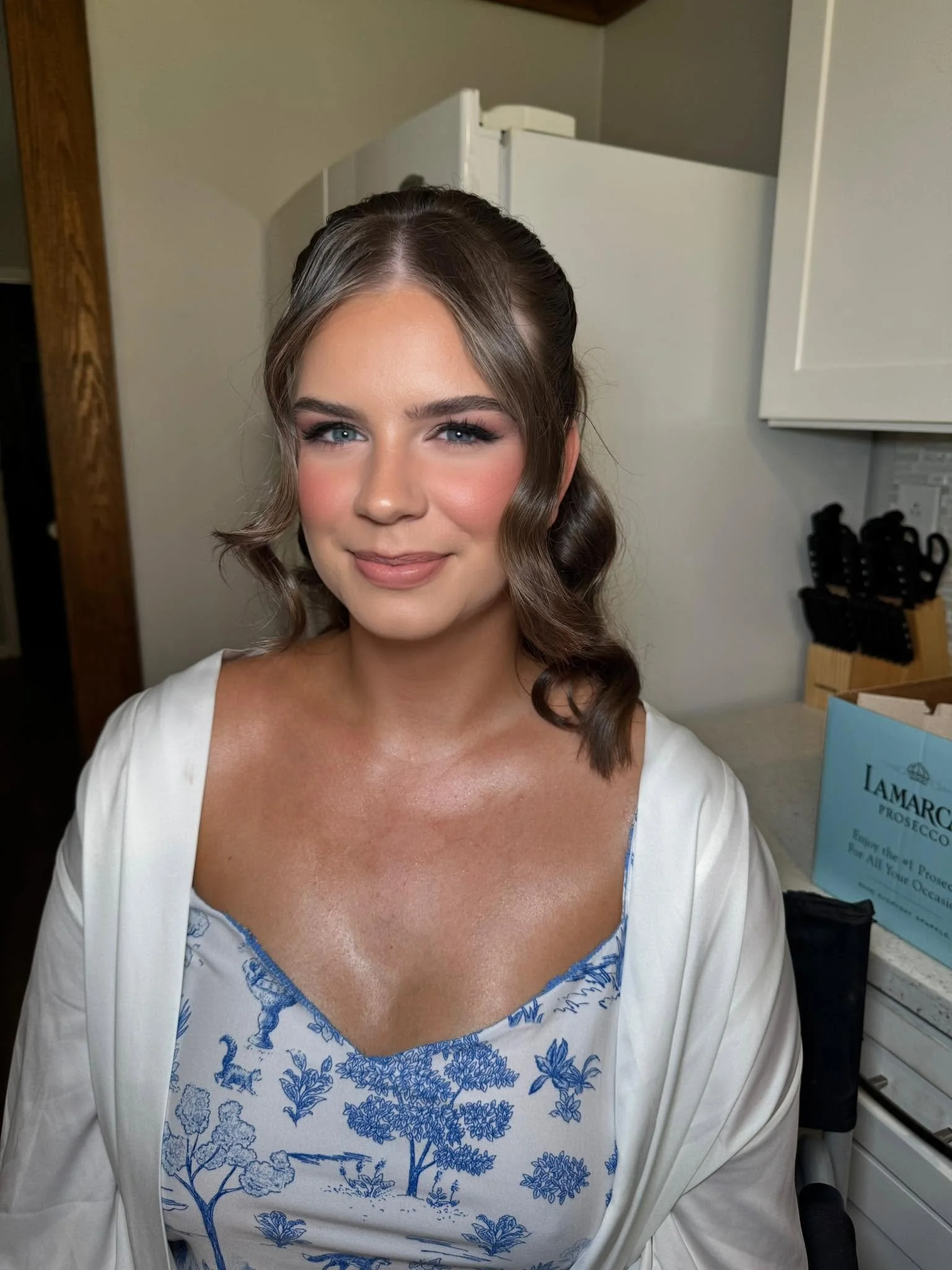 A woman with styled brown hair, blue eyes, and wearing a white dress with blue floral embroidery, standing in a kitchen with white cabinets and knives in the background.