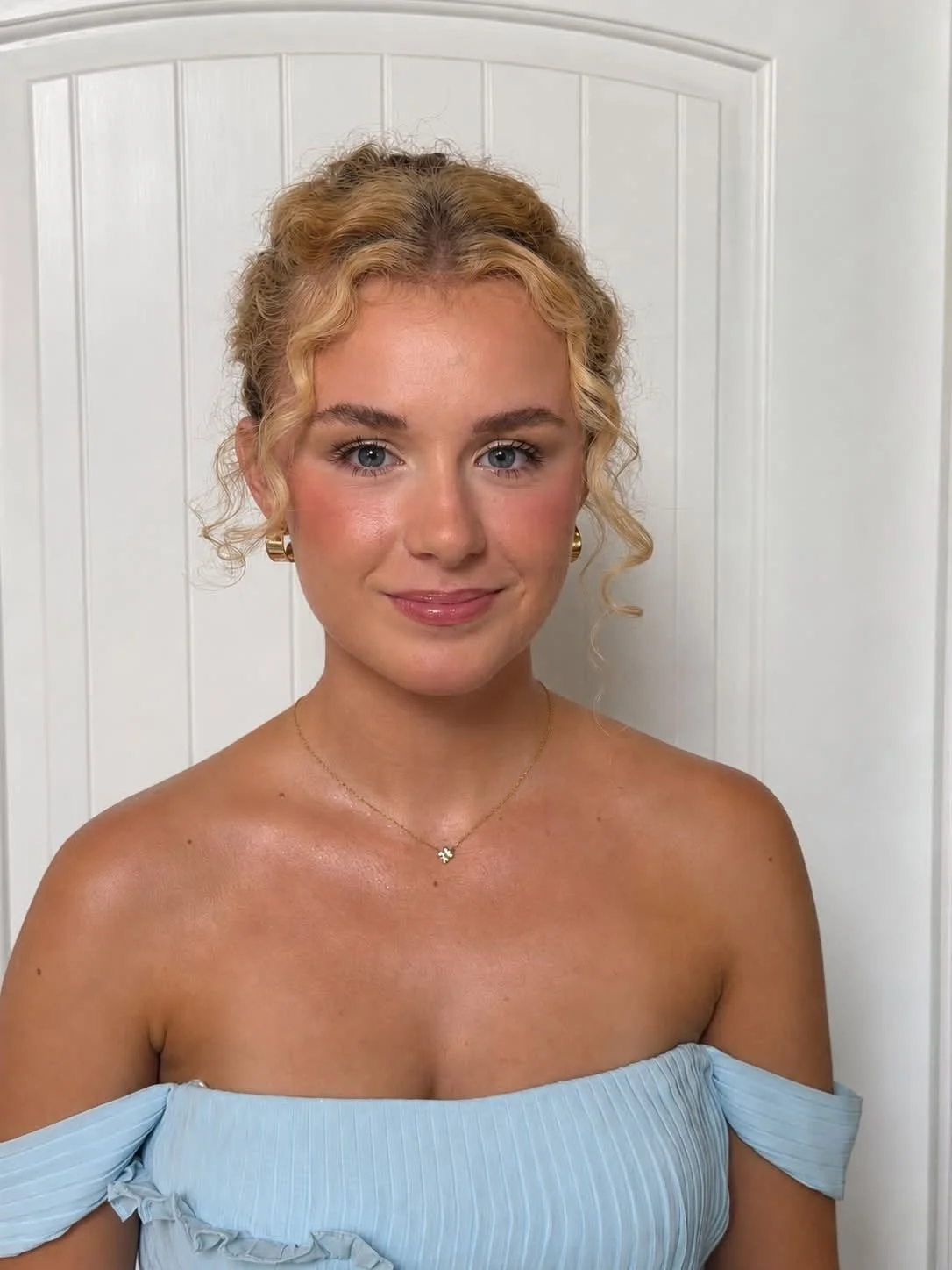 A woman with blonde curly hair and blue eyes, wearing a light blue off-shoulder top and gold jewelry, standing in front of a white paneled door.