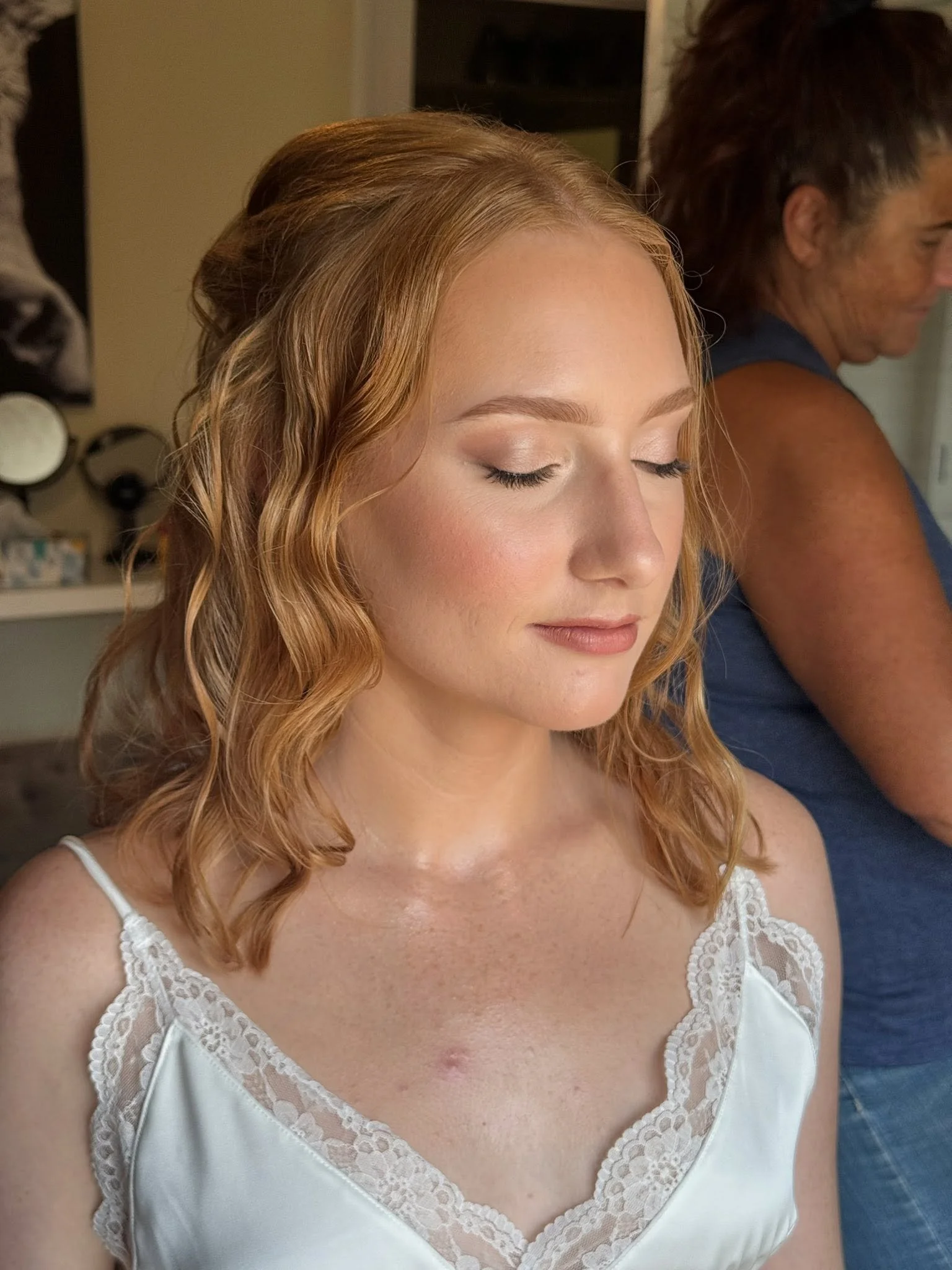A young woman with long, wavy, red hair and closed eyes in a white satin dress with lace details on the straps and neckline, possibly preparing for a special event. Behind her, a woman with short dark hair is partially visible, wearing a sleeveless b