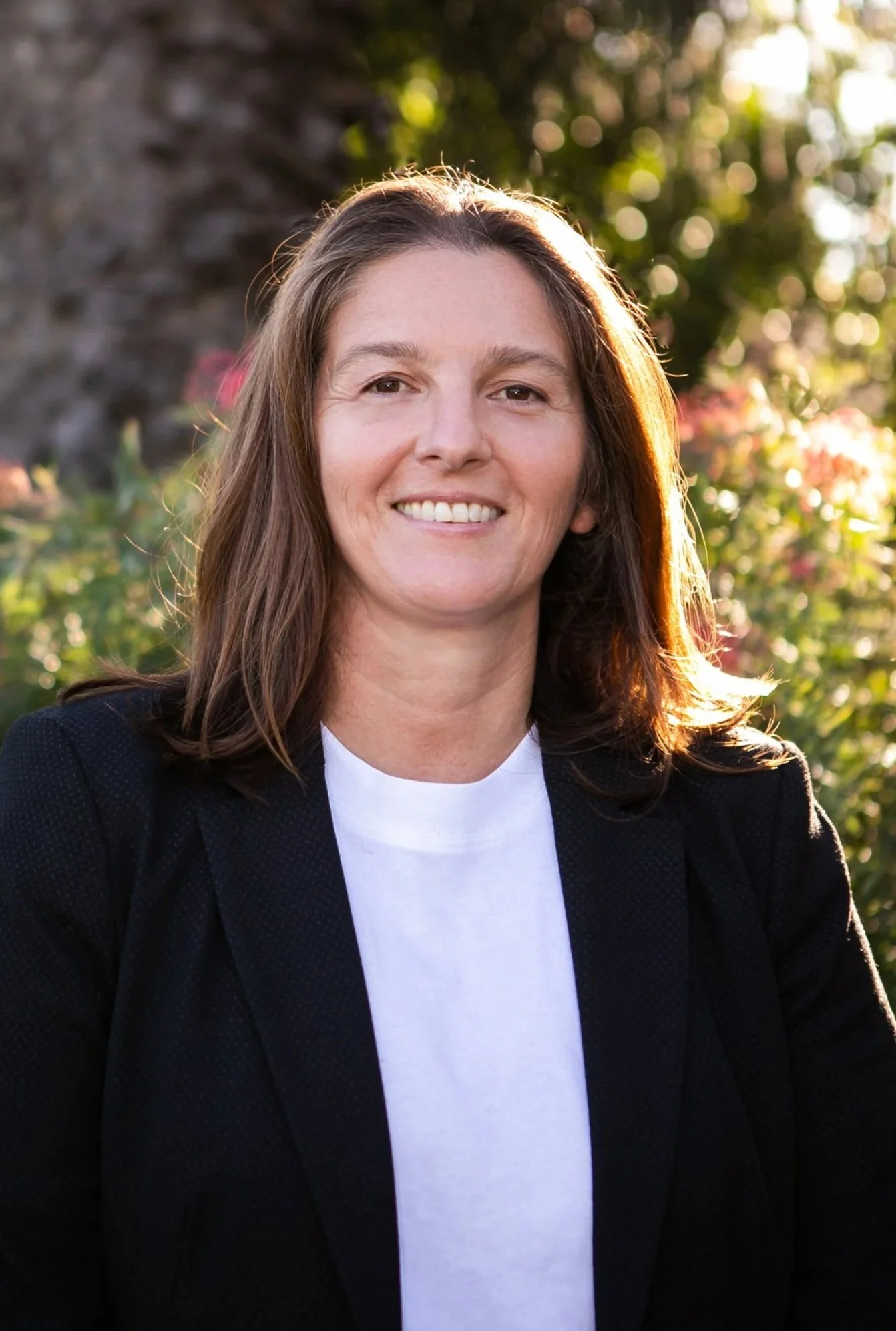 A woman with shoulder-length brown hair, smiling, wearing a black blazer over a white shirt, standing outdoors with a background of sunlight, trees, and foliage.
