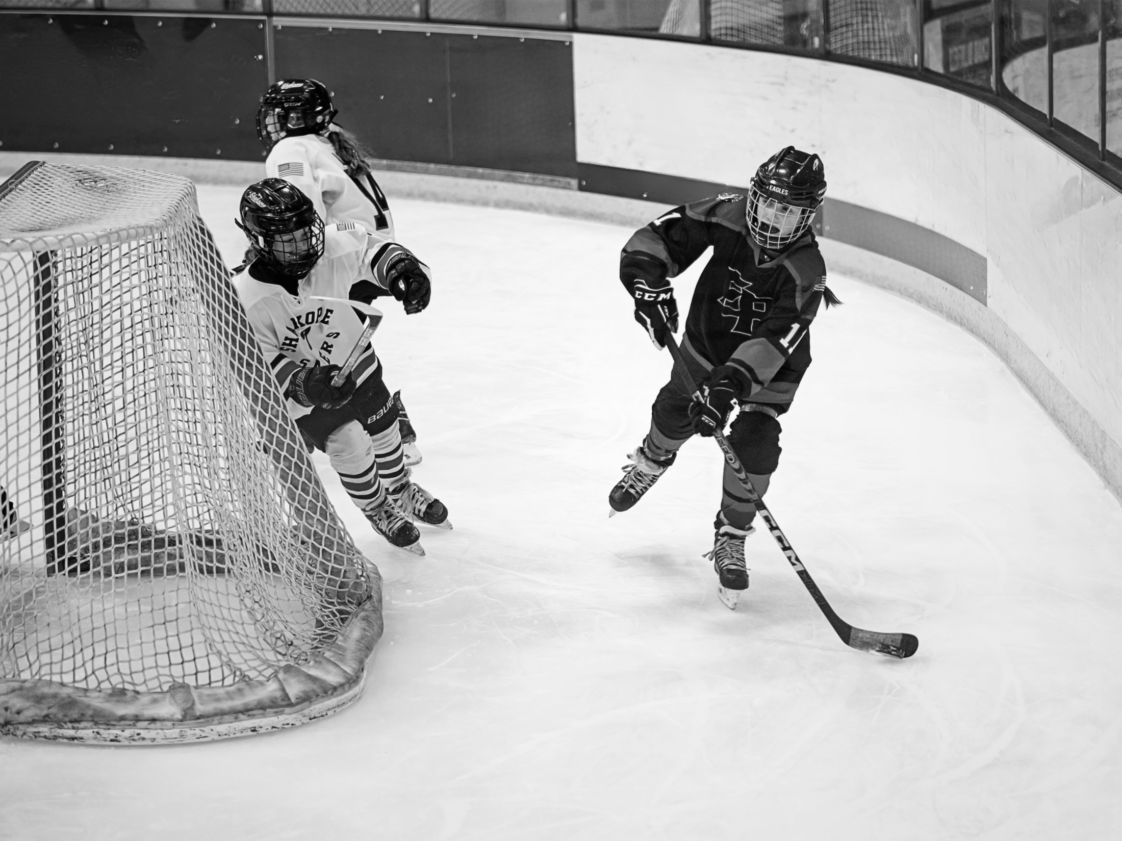 Three young hockey players on the ice near the goal, with one player preparing to shoot or pass the puck as the other two defend or block, inside an ice hockey rink.