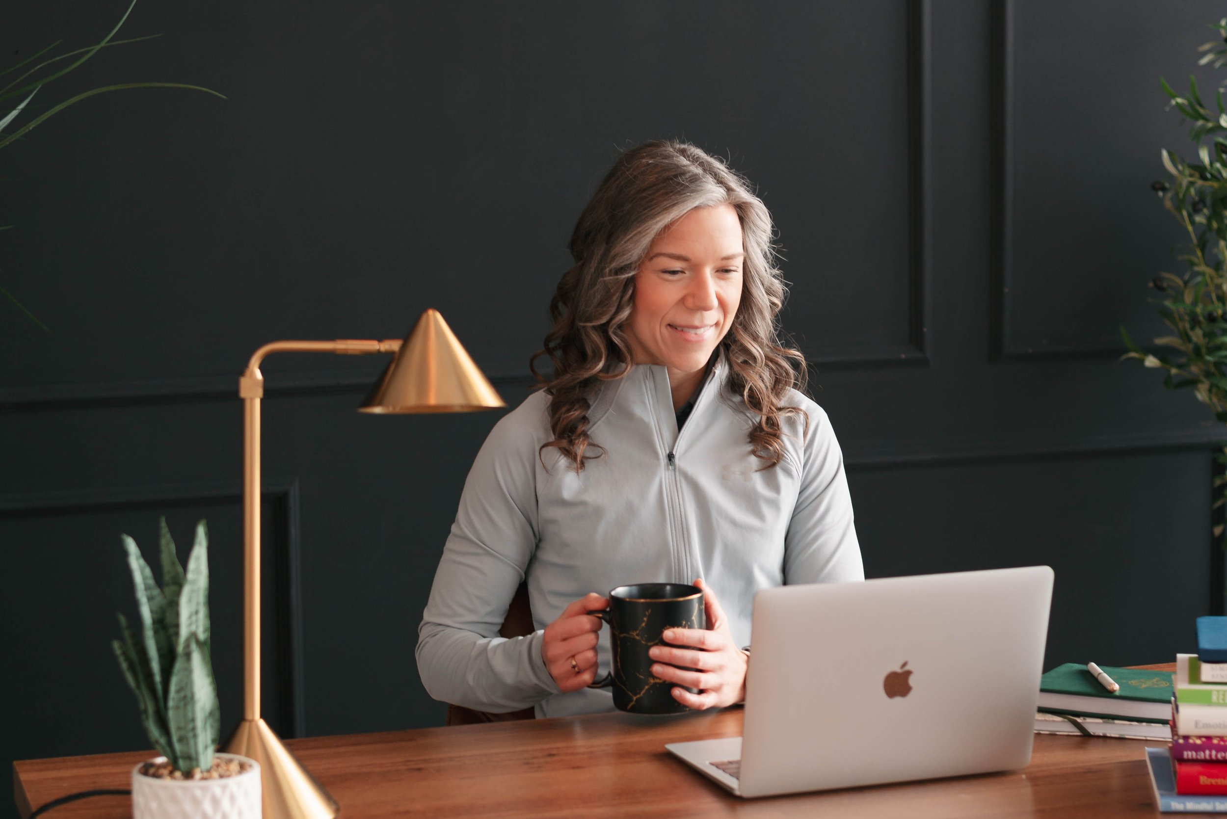 A woman with wavy hairstyles, wearing a gray jacket, is sitting at a desk holding a black mug with a gold marbled design. She is working on a laptop, with a white laptop and some stacked books on her desk. The background features a dark wall with green plants and a gold desk lamp.