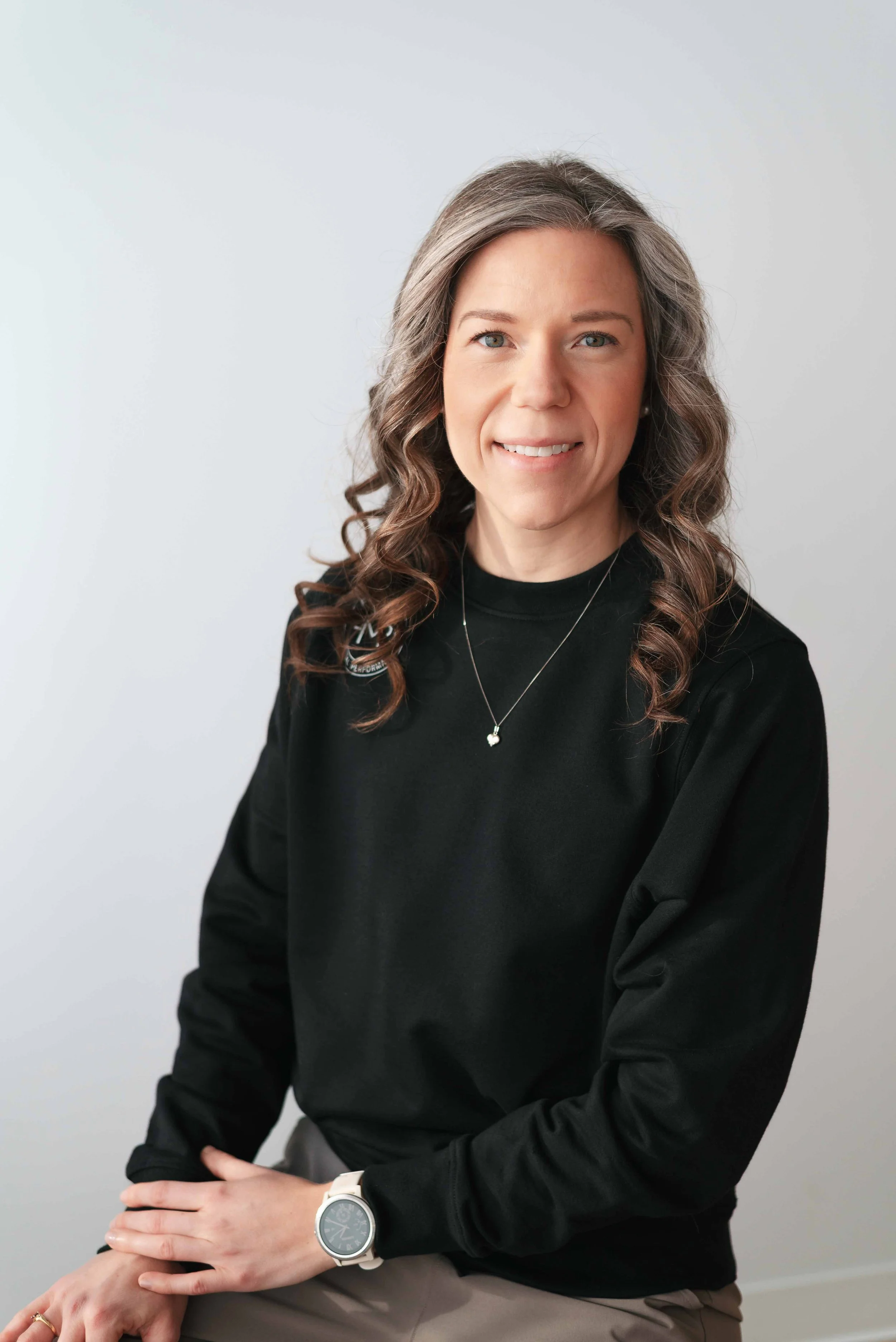 A woman with long, curly gray hair and blue eyes, smiling, wearing a black long-sleeve shirt, beige pants, a white watch, and a necklace with a heart pendant, sitting against a plain light gray background.