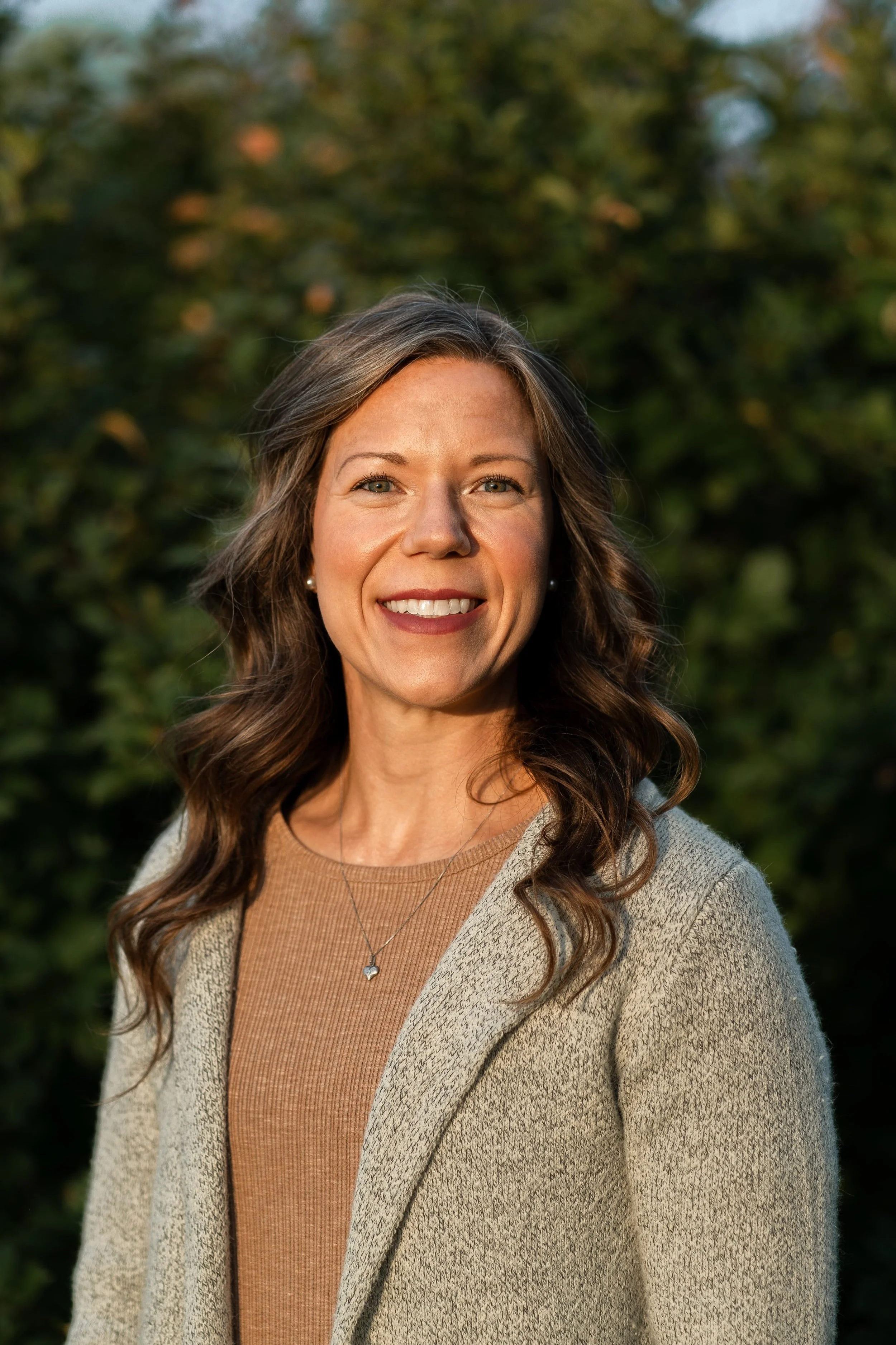 A woman with wavy brown hair and blue eyes smiling outdoors in front of green trees, wearing a beige cardigan, a light brown top, and a heart-shaped necklace.