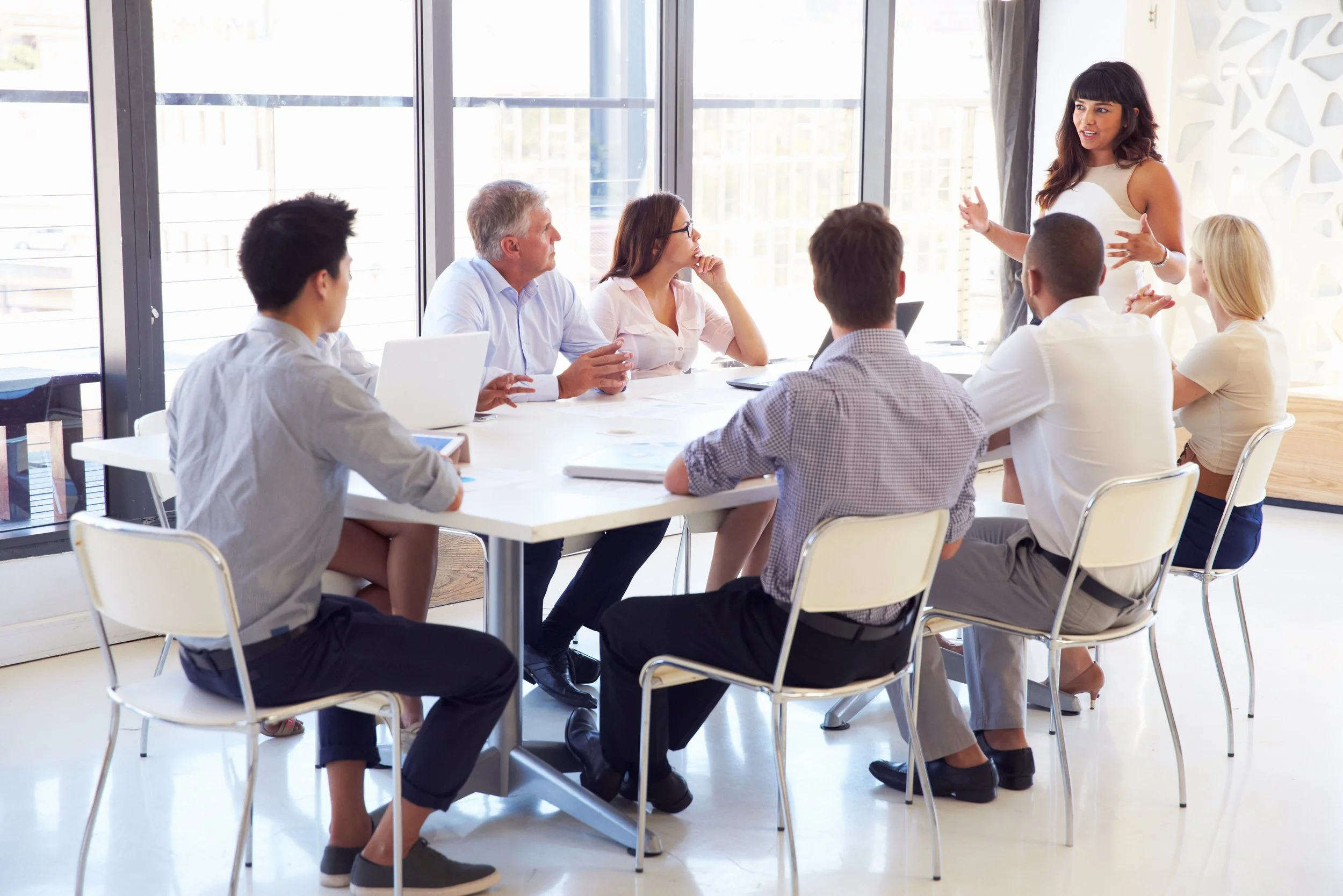 Team of people sitting around table listening to woman leading discussion