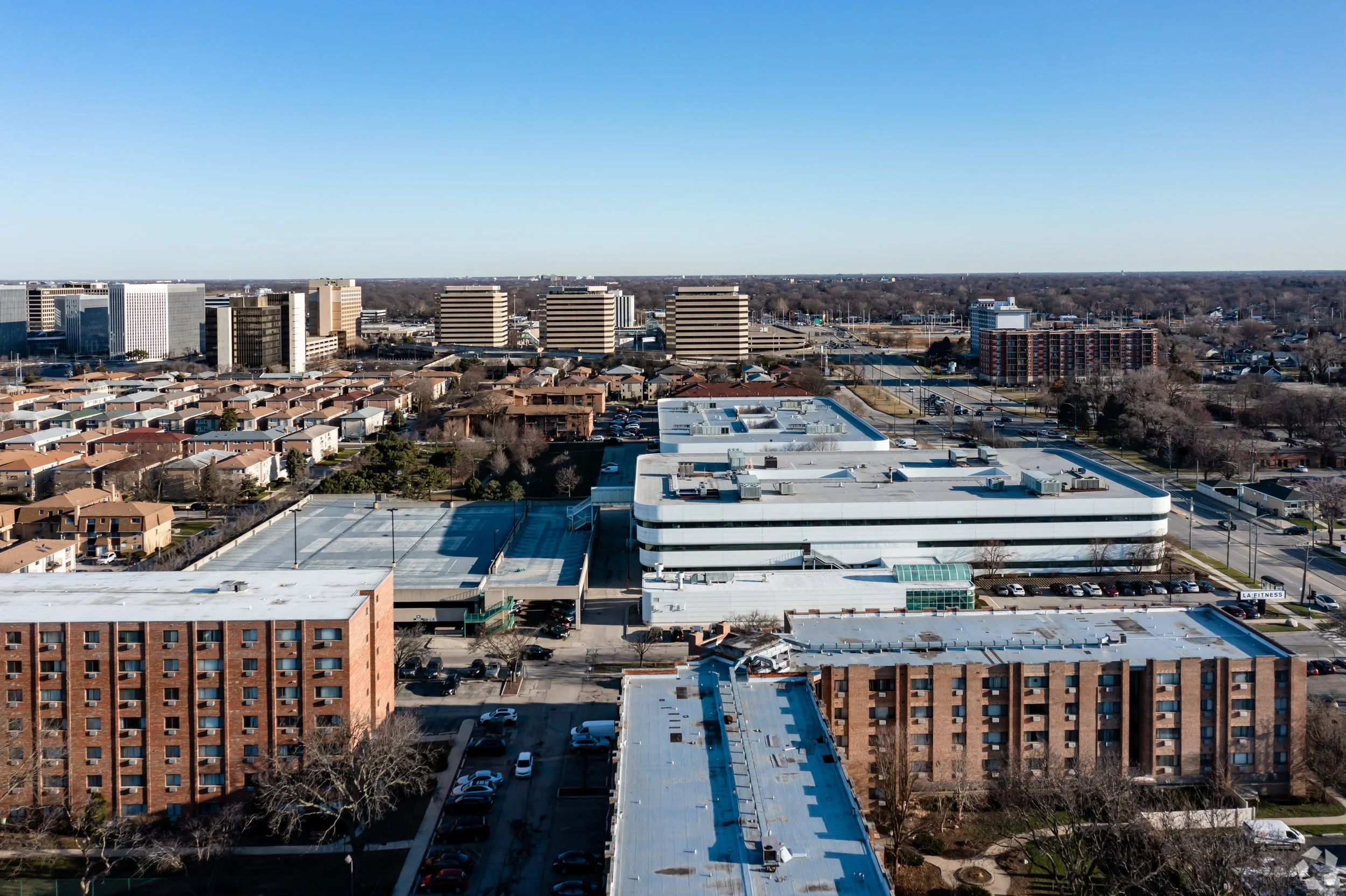 Aerial view of suburban area with commercial buildings and large parking lots, surrounded by residential neighborhoods and expansive flat terrain.