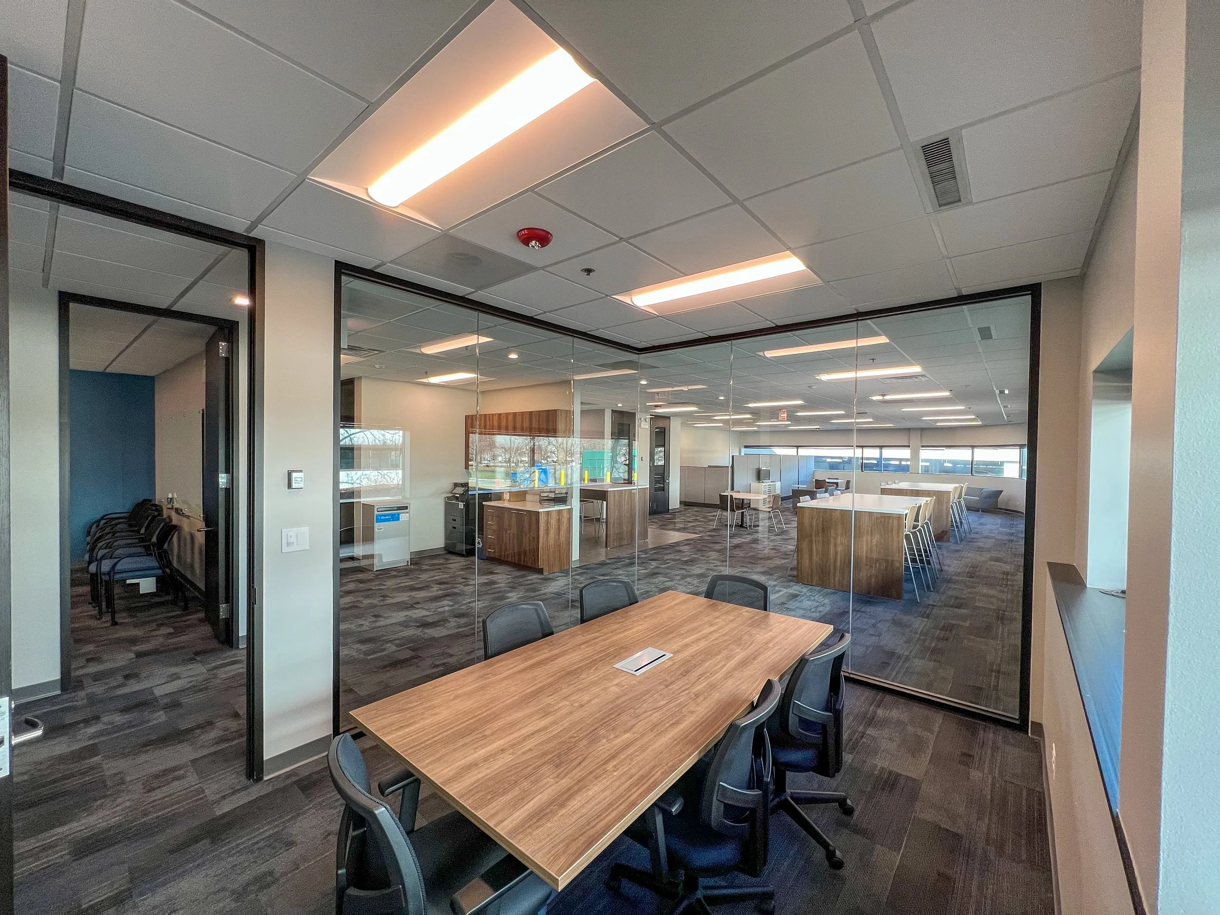 Modern office space with a glass-walled meeting room, several chairs, tables, and workstations. The office has a clean, contemporary design with a drop ceiling and dark carpeting.