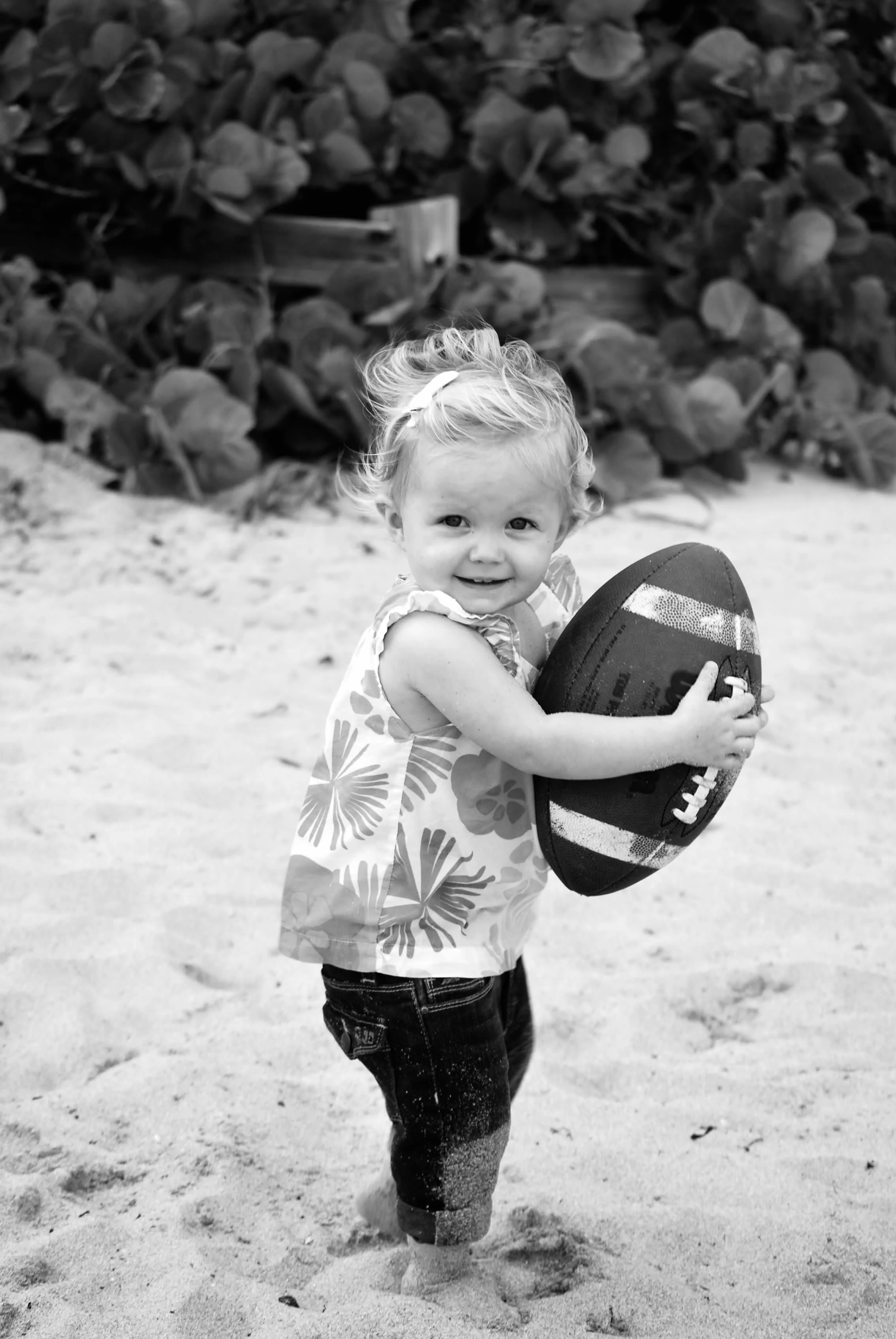 A young girl smiling and holding a rugby ball on a sandy beach, with large leafy plants in the background.