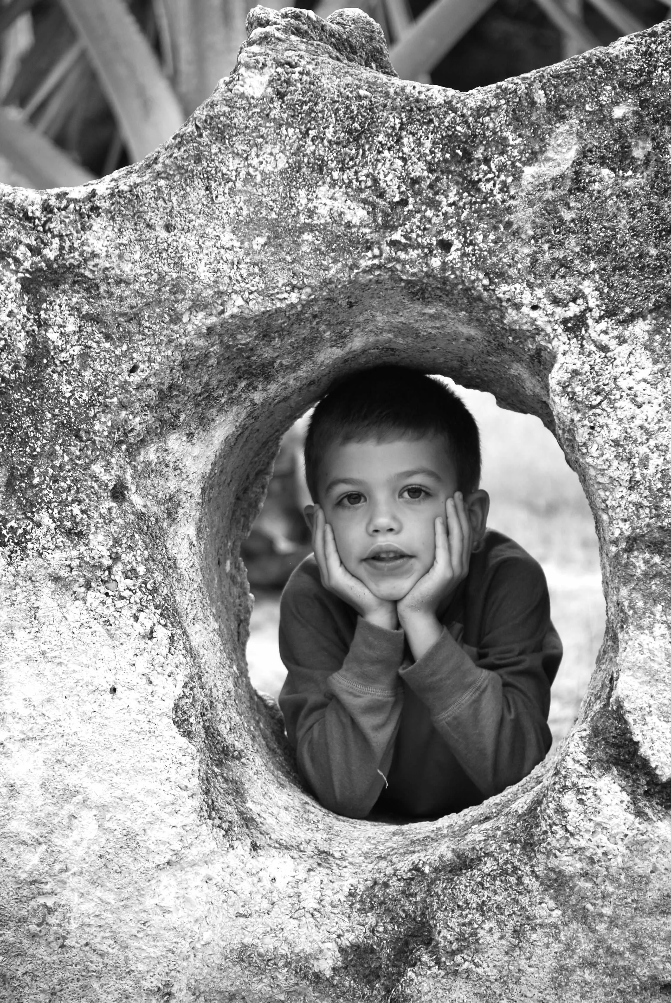 A young boy with short hair resting his chin in his hands, looking through a large, textured, circular hole in a rock or sculpture.