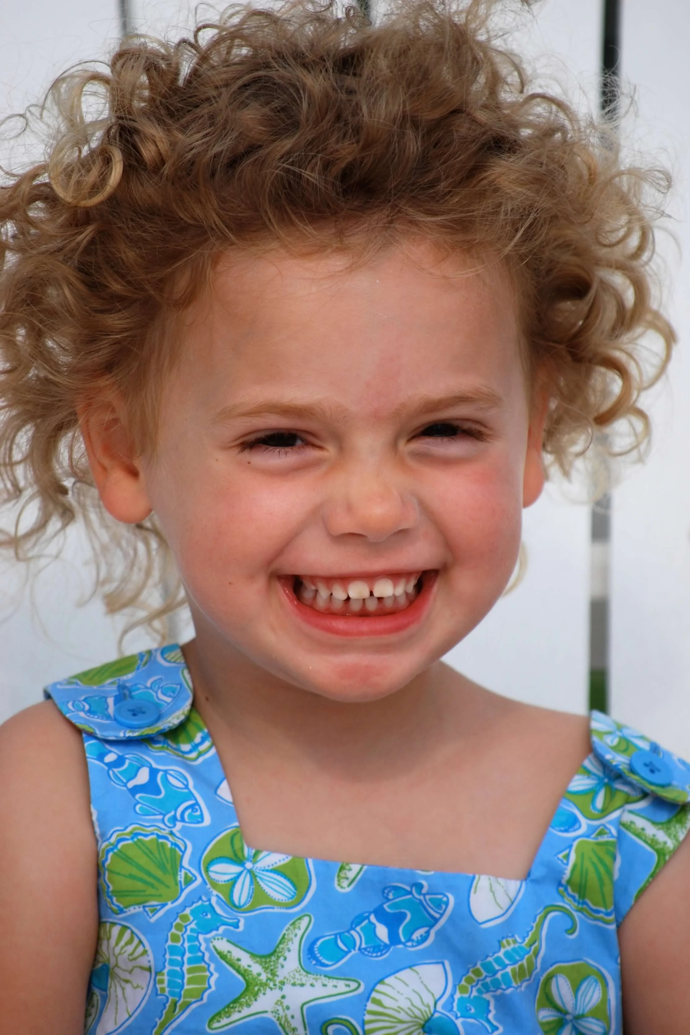 A young girl with curly blonde hair smiling and showing her teeth, wearing a blue dress with seashells, starfish, and fish prints.