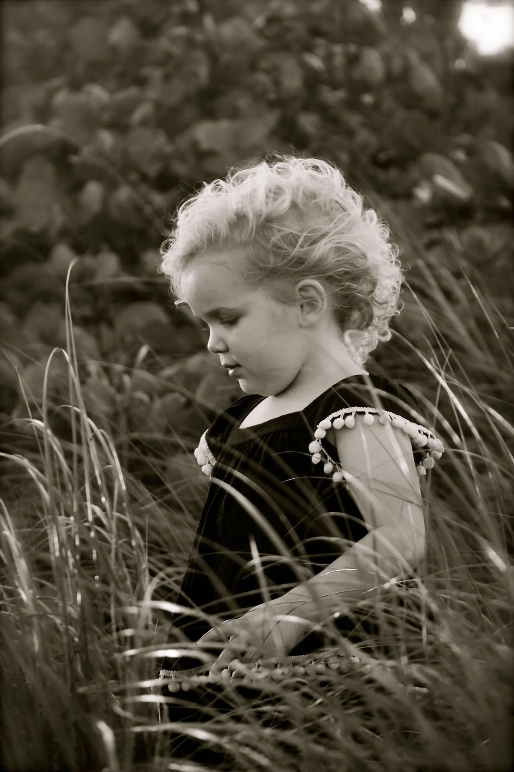A young girl with curly blonde hair standing in tall grass, wearing a dark dress with pom-pom embellishments on the sleeves, looking down thoughtfully in a natural outdoor setting.