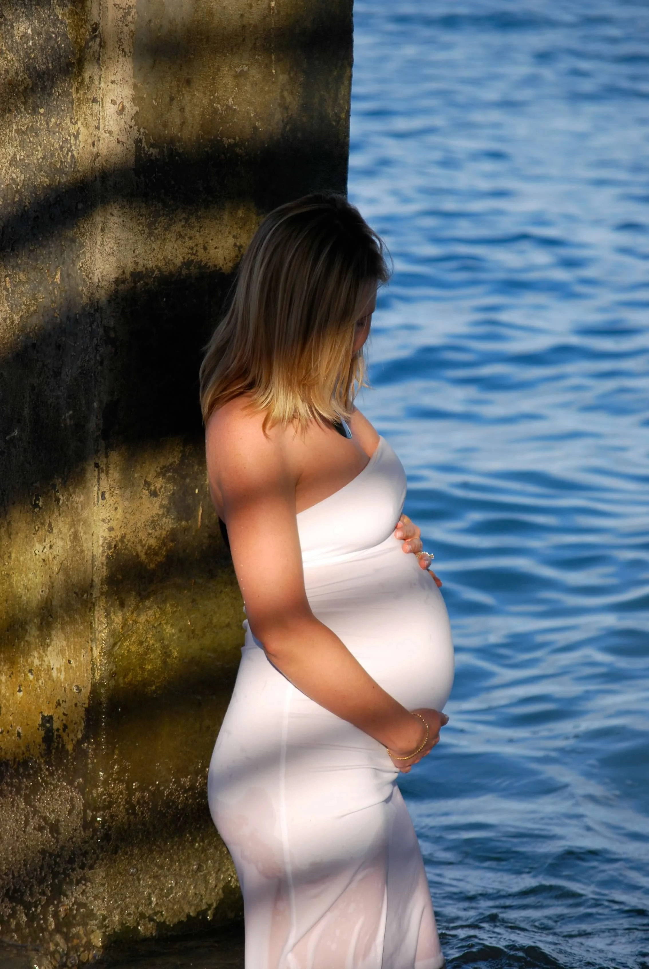 Pregnant woman in a white dress leaning against a mossy wall near the water at sunset.