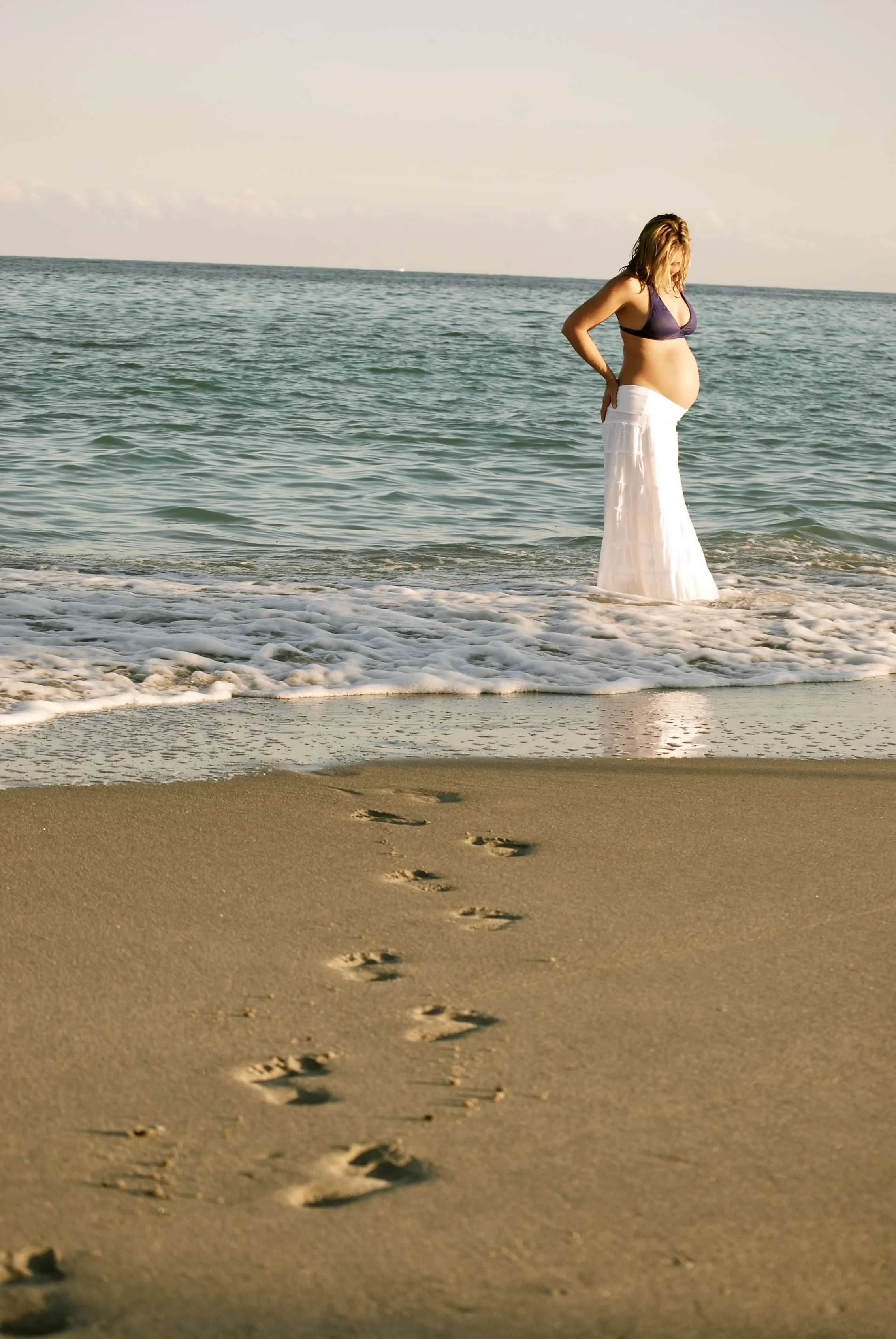 A pregnant woman in a dark bikini top and white skirt standing in the shallow surf at the beach, with footprints leading to her on the sand, during sunset or sunrise.