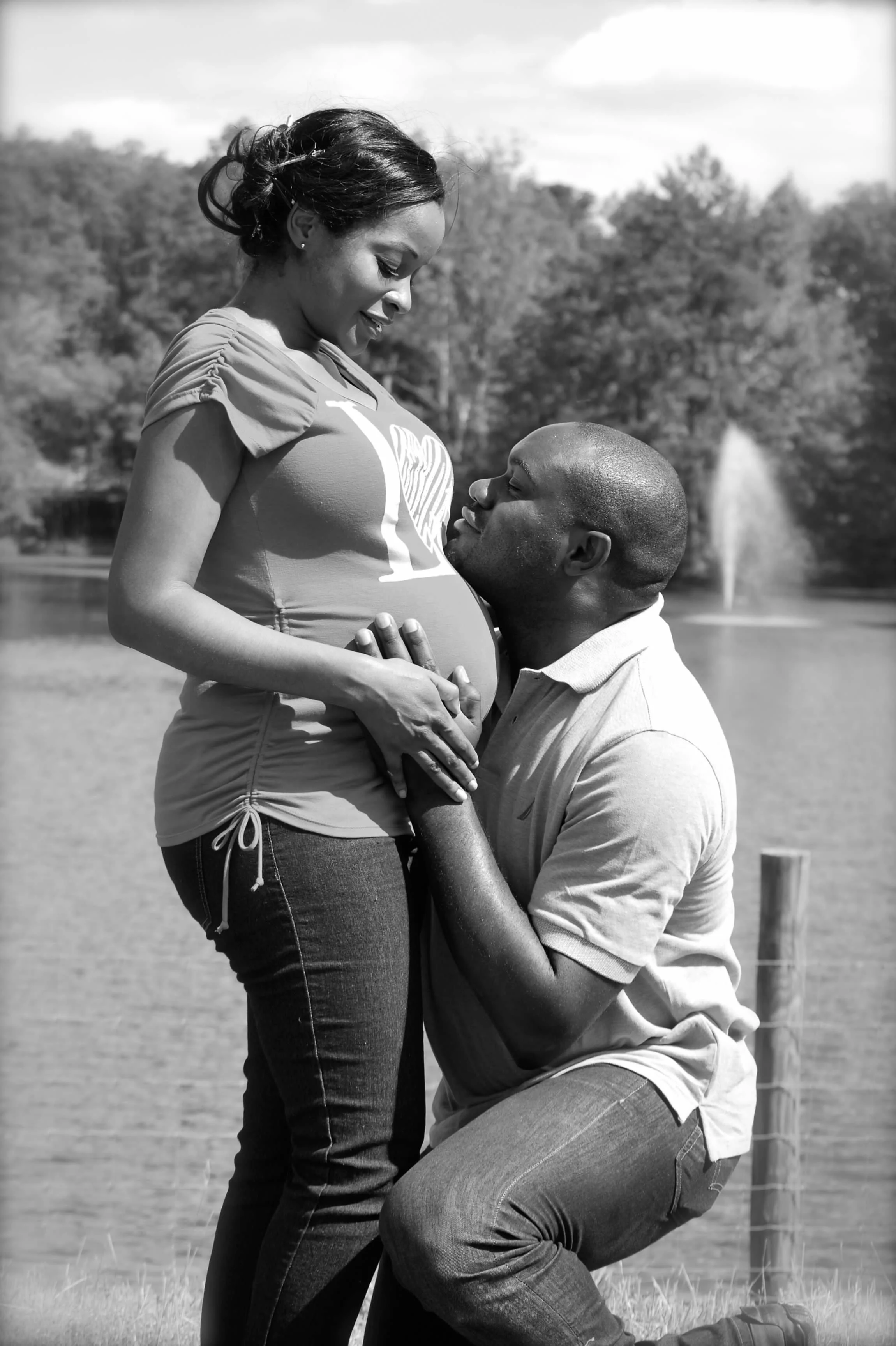 A romantic outdoor scene in black and white between a pregnant woman and her partner. The man is kneeling and gazing into the woman’s eyes, placing his hand on her belly. The woman is standing, smiling down at him, with her hand on his shoulder. Ther