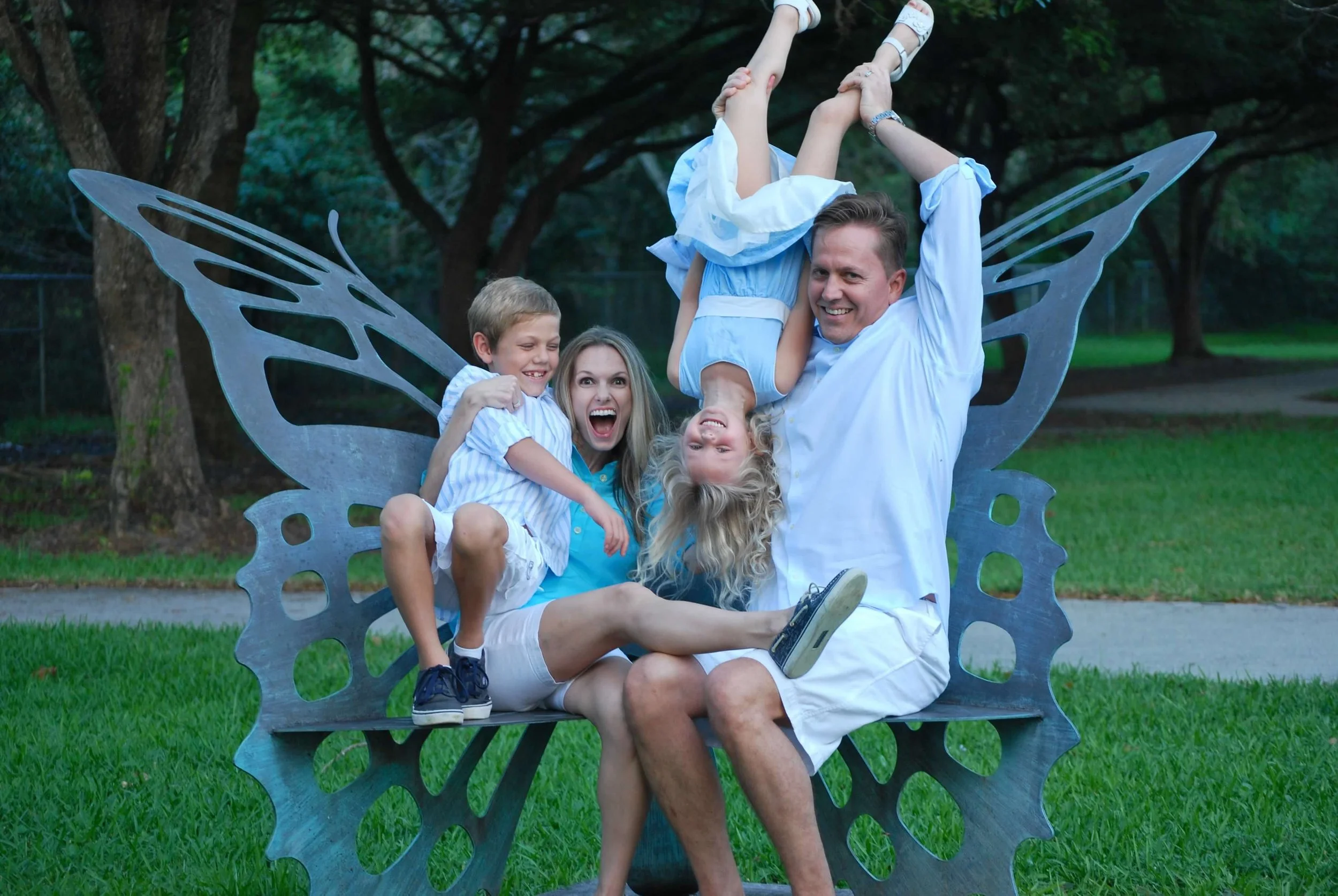 Family of four with butterfly wings, playing and smiling on a park bench with green trees in the background.
