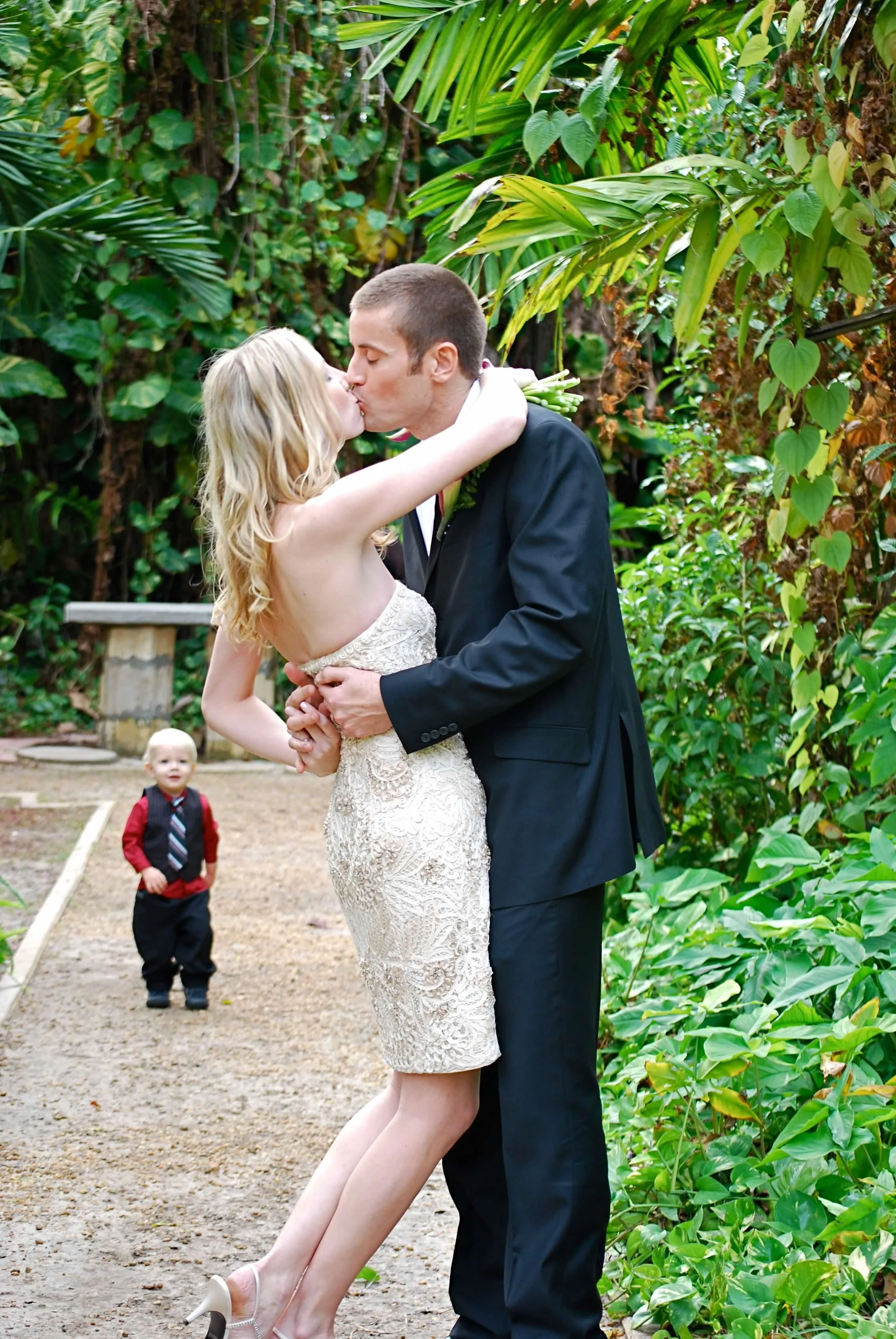 A couple in wedding attire sharing a kiss in a lush, green outdoor setting, with a small child in formal clothing watching in the background.