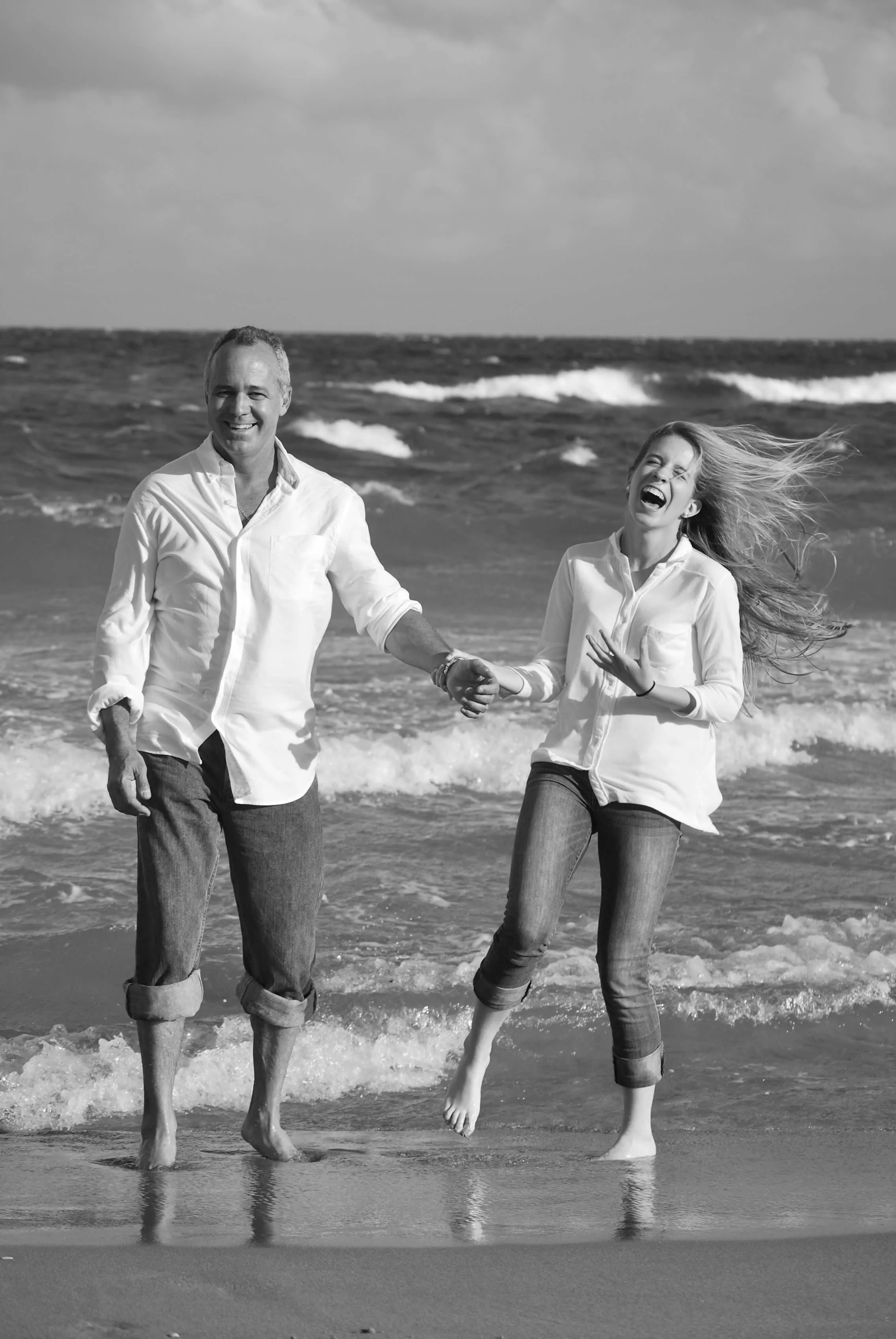 A man and woman holding hands and laughing on the beach with waves in the background.