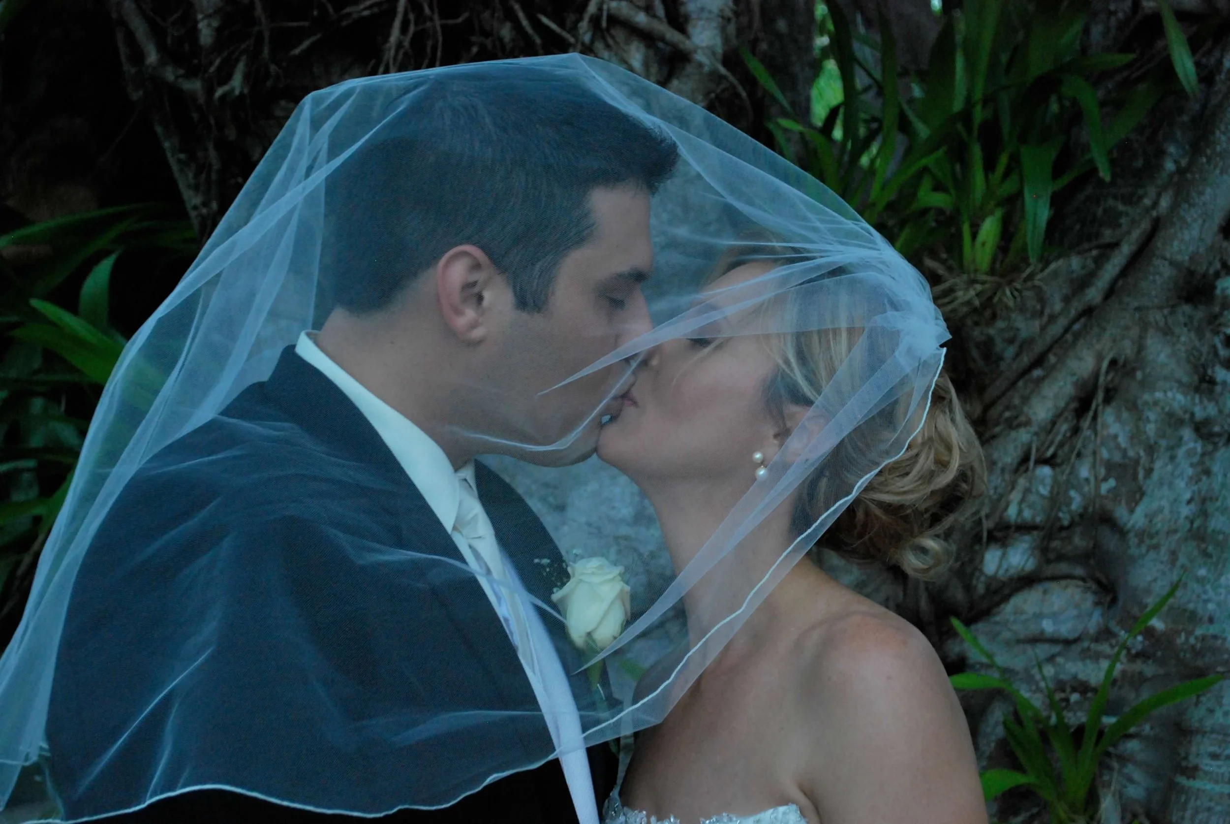 A newlywed couple sharing a kiss under a wedding veil in a natural outdoor setting with trees and greenery in the background.