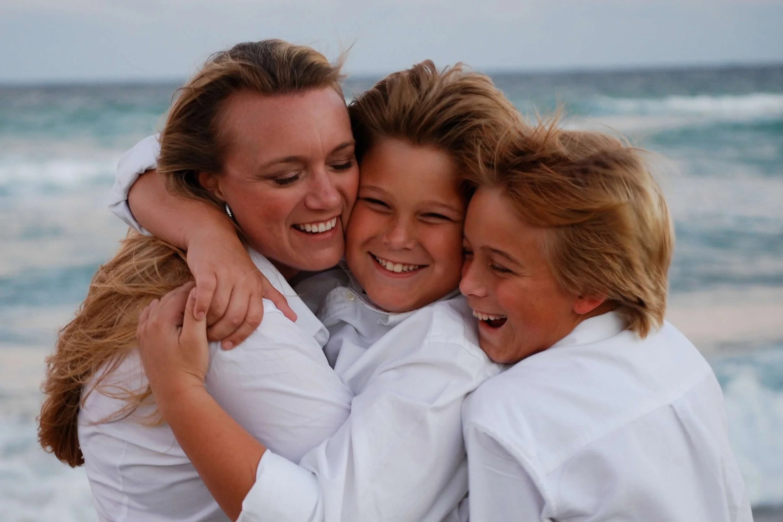 Three women in white shirts hug each other on a beach with ocean waves in the background, smiling and laughing.