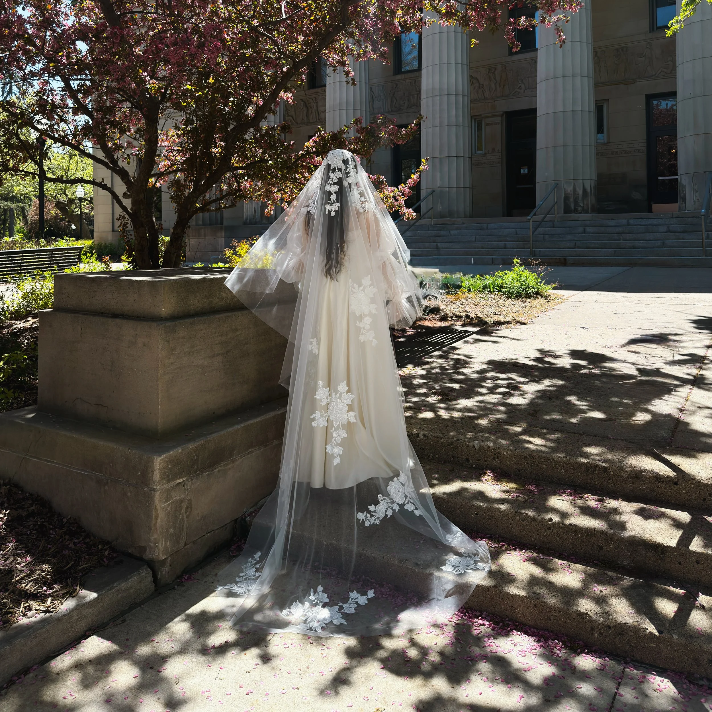 The photo shows a bride standing on the stairs with her face and body turned away. The full veil is visible with large floral lace on it