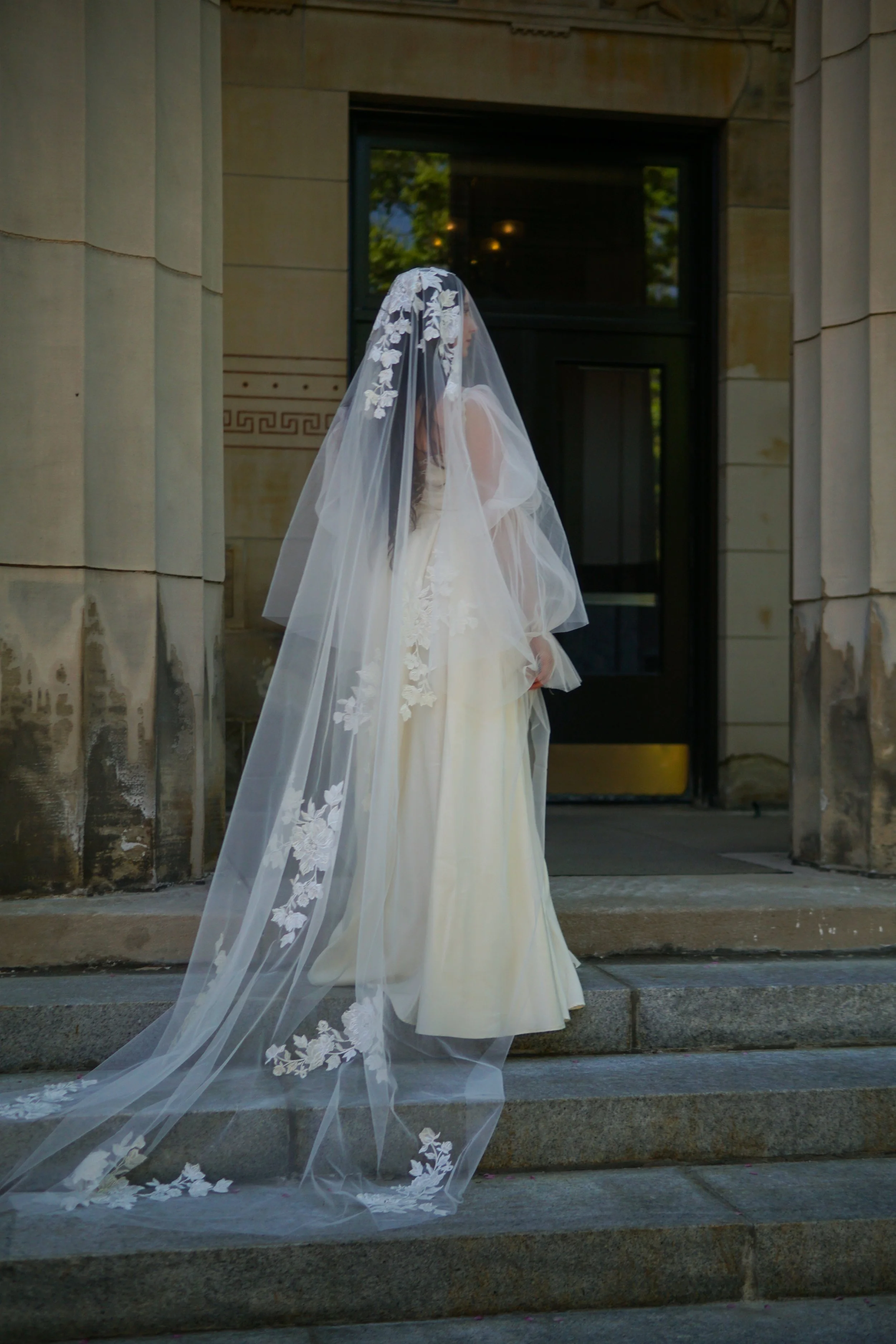 The photo shows a bride standing on the stairs with her face and body turned away. The full veil is visible with large floral lace t entire veil and large floral lace on the head area