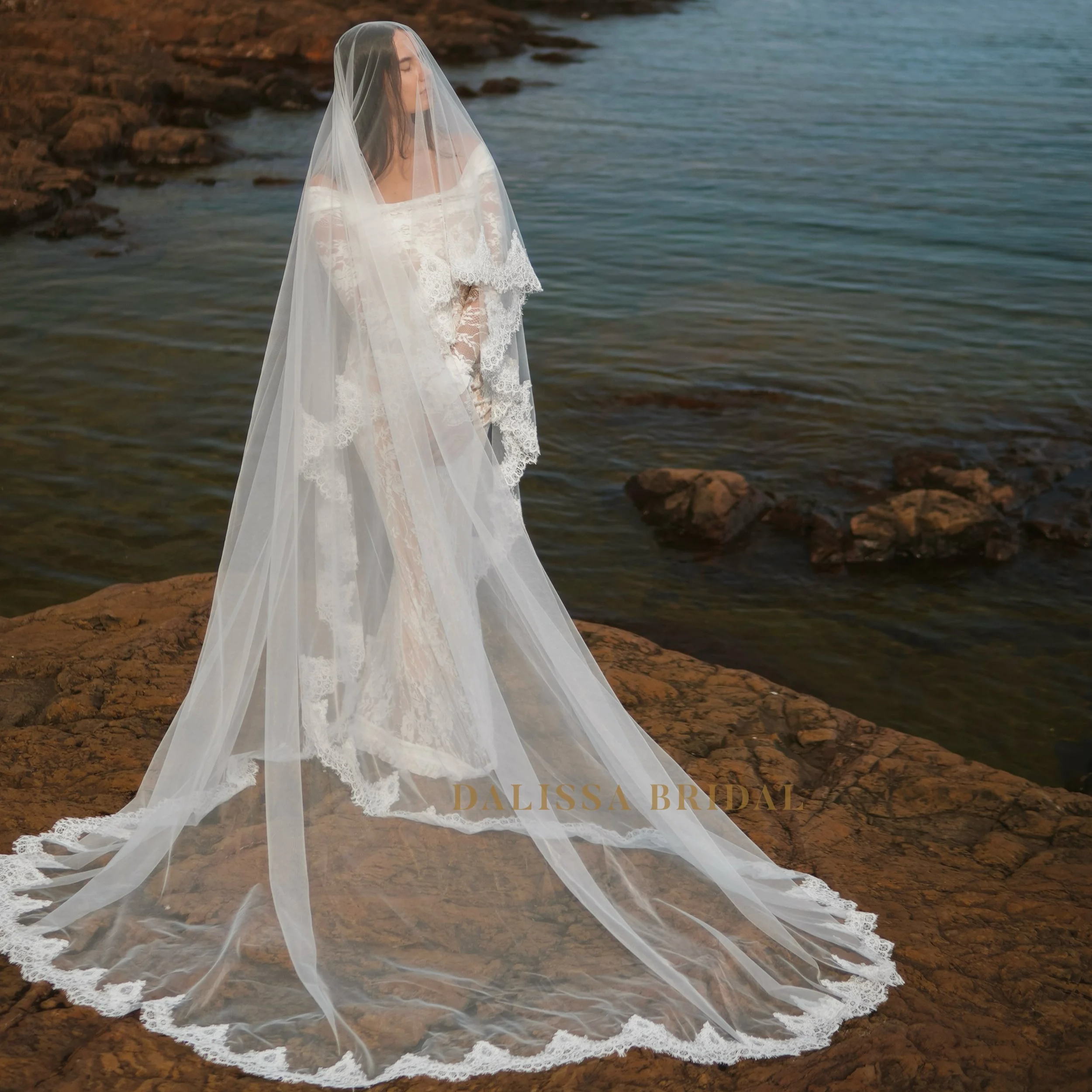 Bride wearing a sheer cathedral-length wedding veil with delicate Chantilly lace trim, standing on rocky shoreline overlooking the ocean.