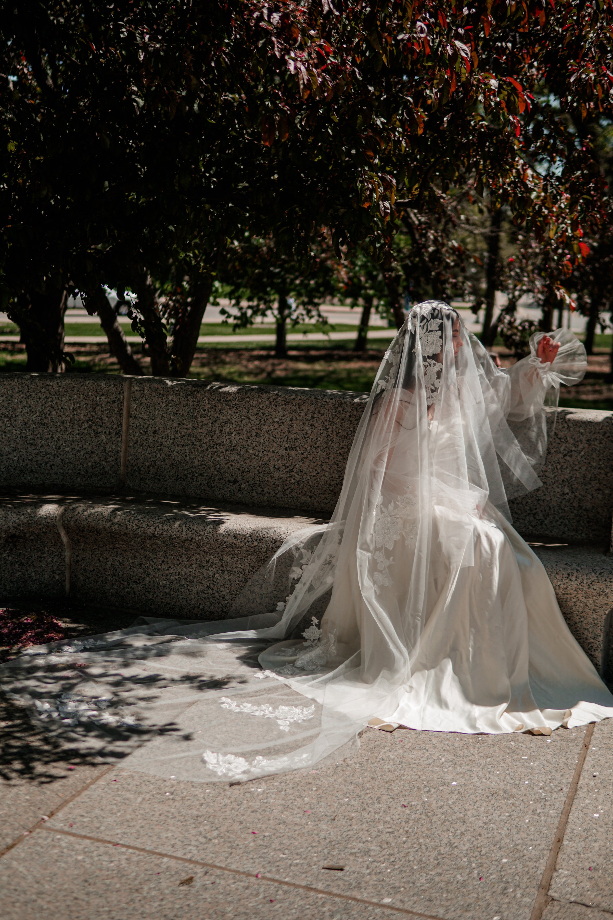 Bride seating in the garden in blusher floral lace veil in cathedral length. The lace is covering face partially from the side.