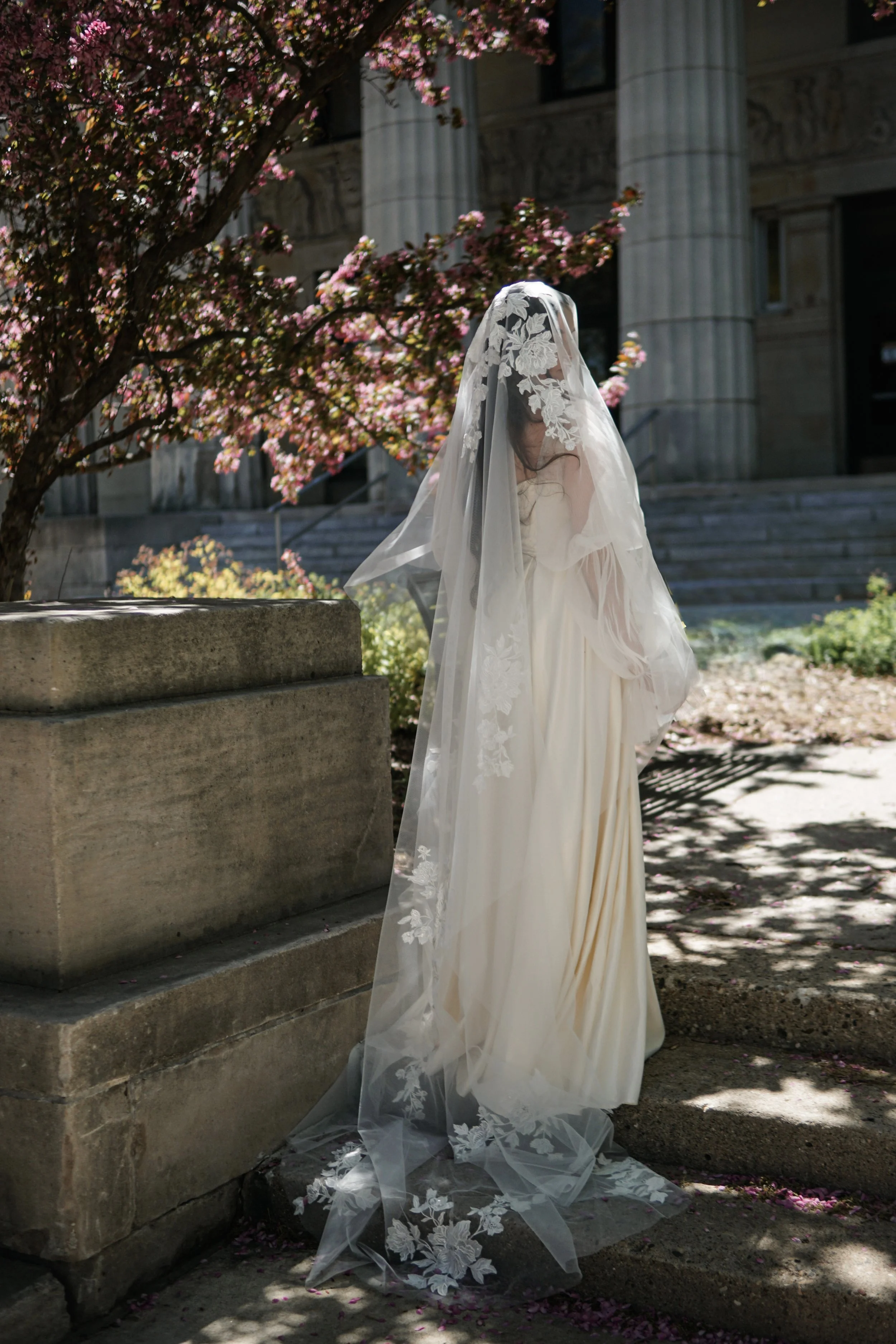 The photo shows a bride standing on the stairs with her face and body turned away. The full veil is visible with large floral lace on it