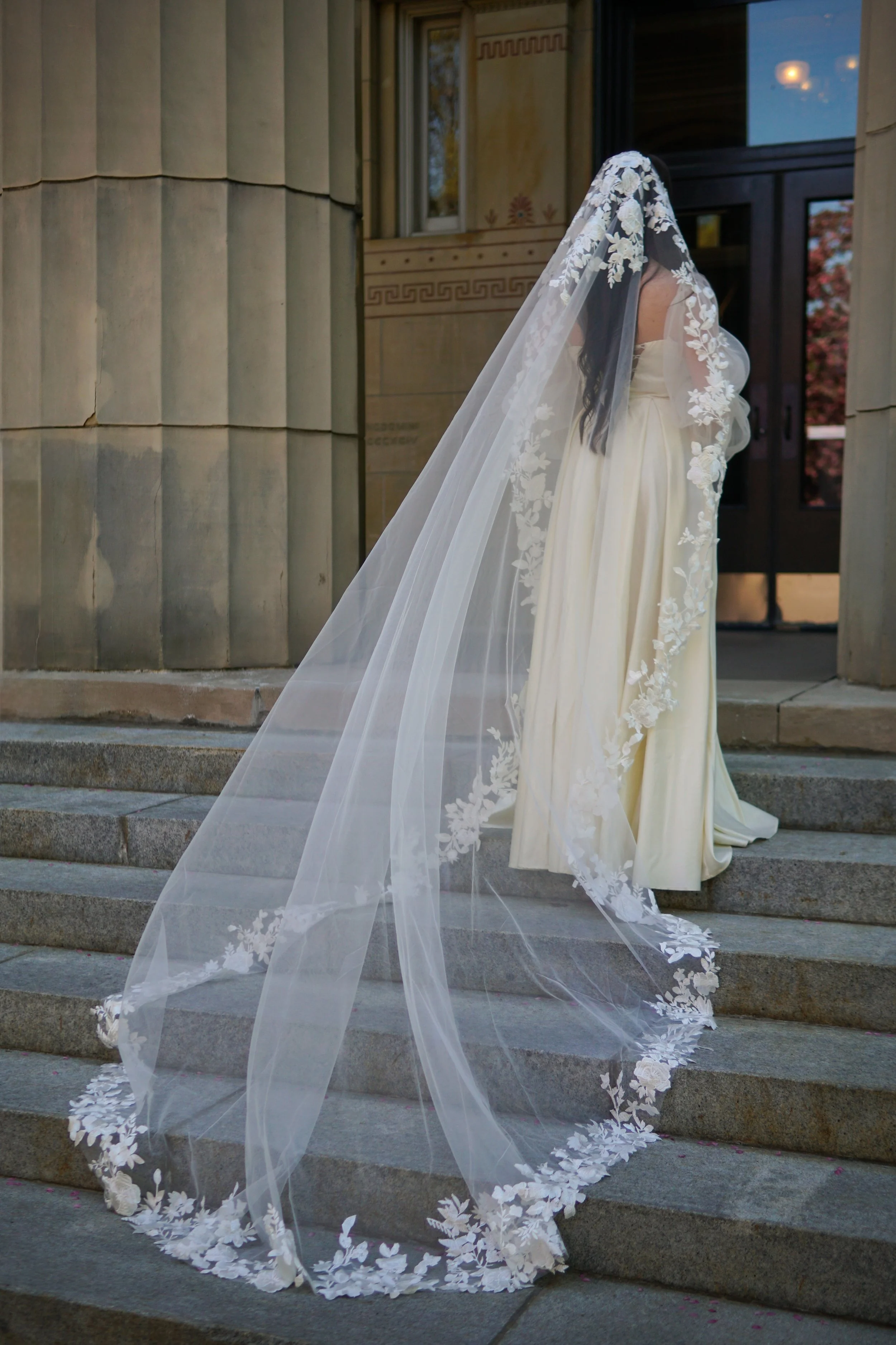 The soft ivory tulle veil gracefully drapes over the bride and cascades into a dramatic train, with dimensional floral lace framing the head and edging the veil.