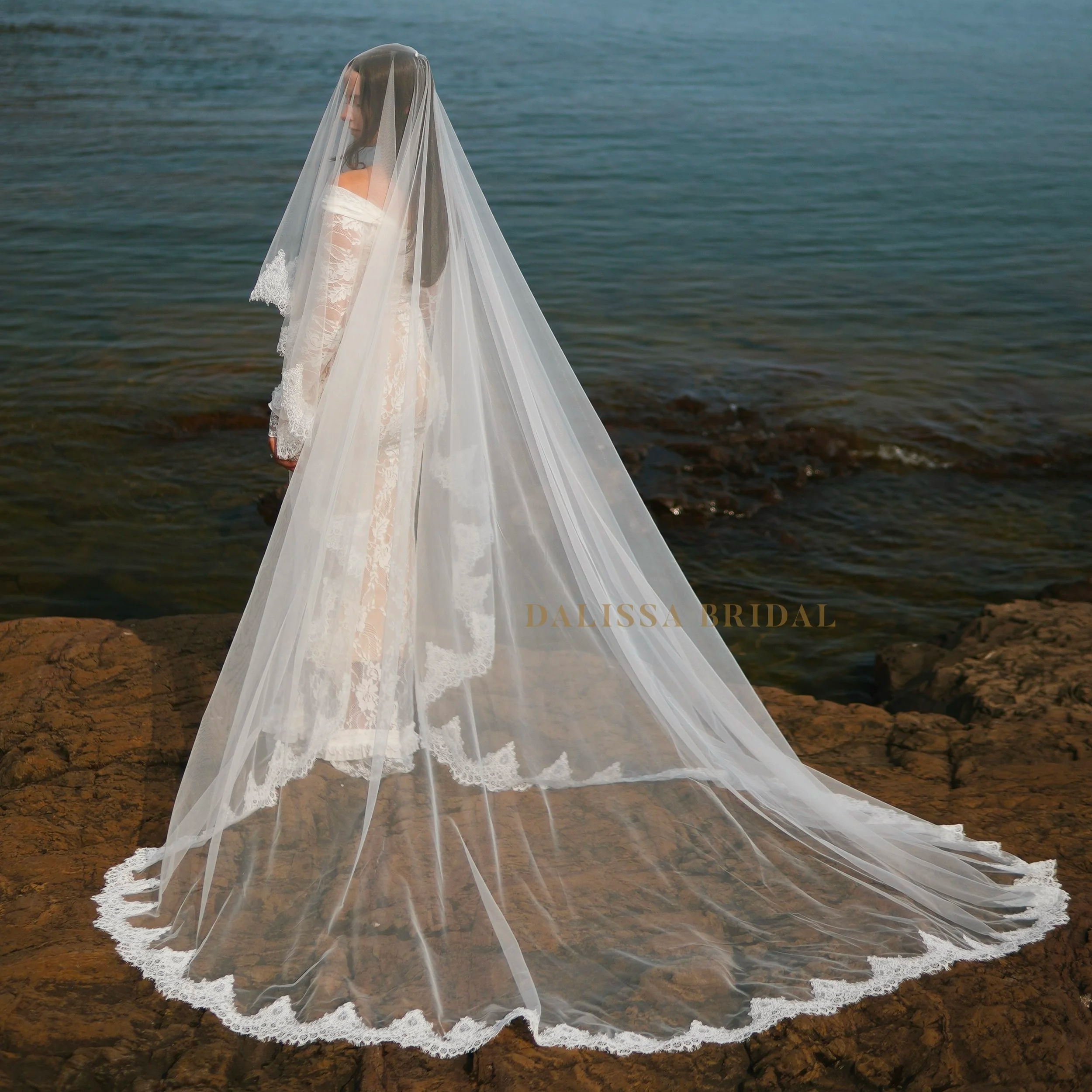 Bride wearing a sheer cathedral-length wedding veil with delicate Chantilly lace trim, standing on rocky shoreline overlooking the ocean.