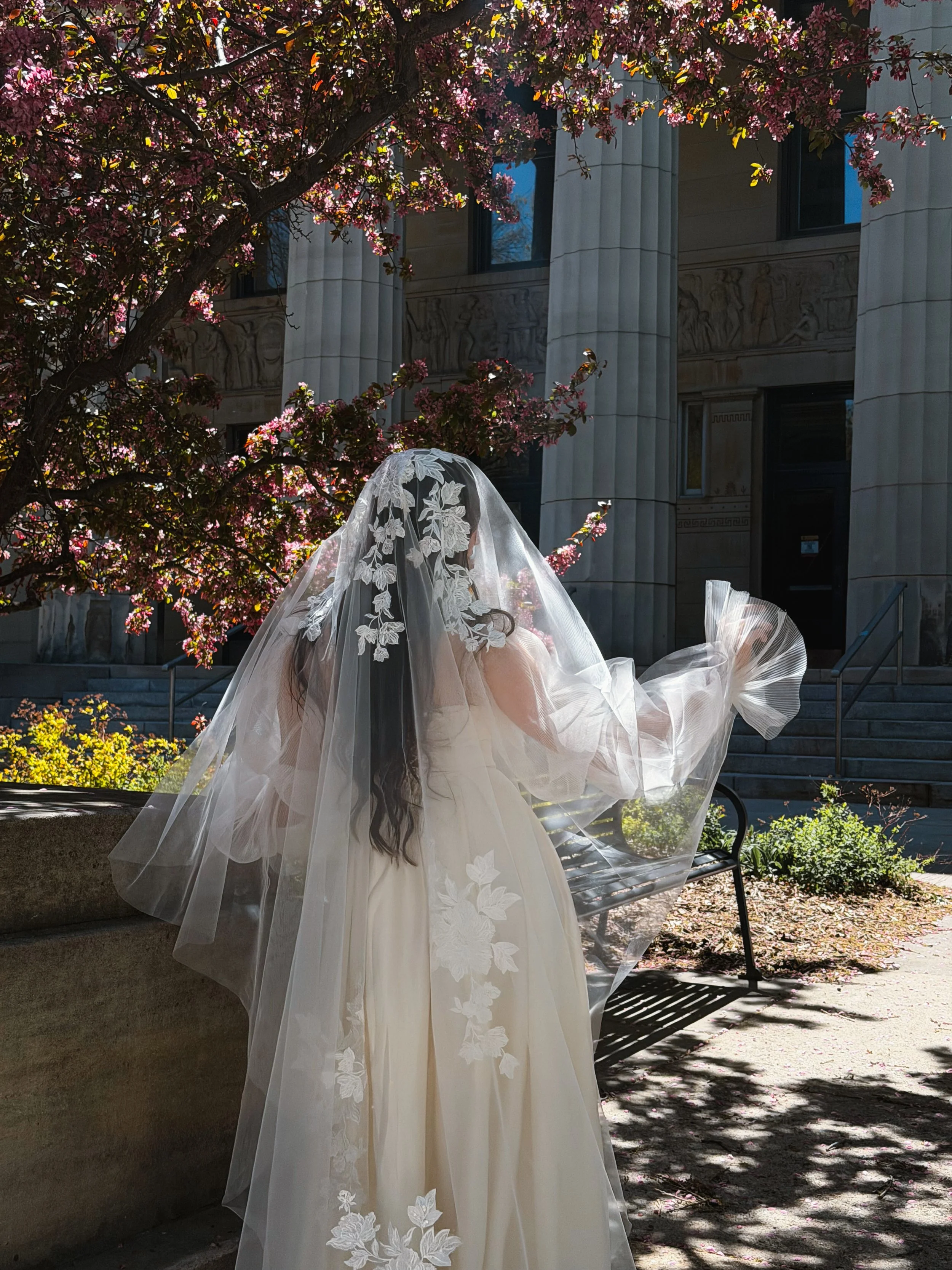 The photo shows a bride standing on the stairs with her face and body turned away. The half veil is visible with large floral lace on it