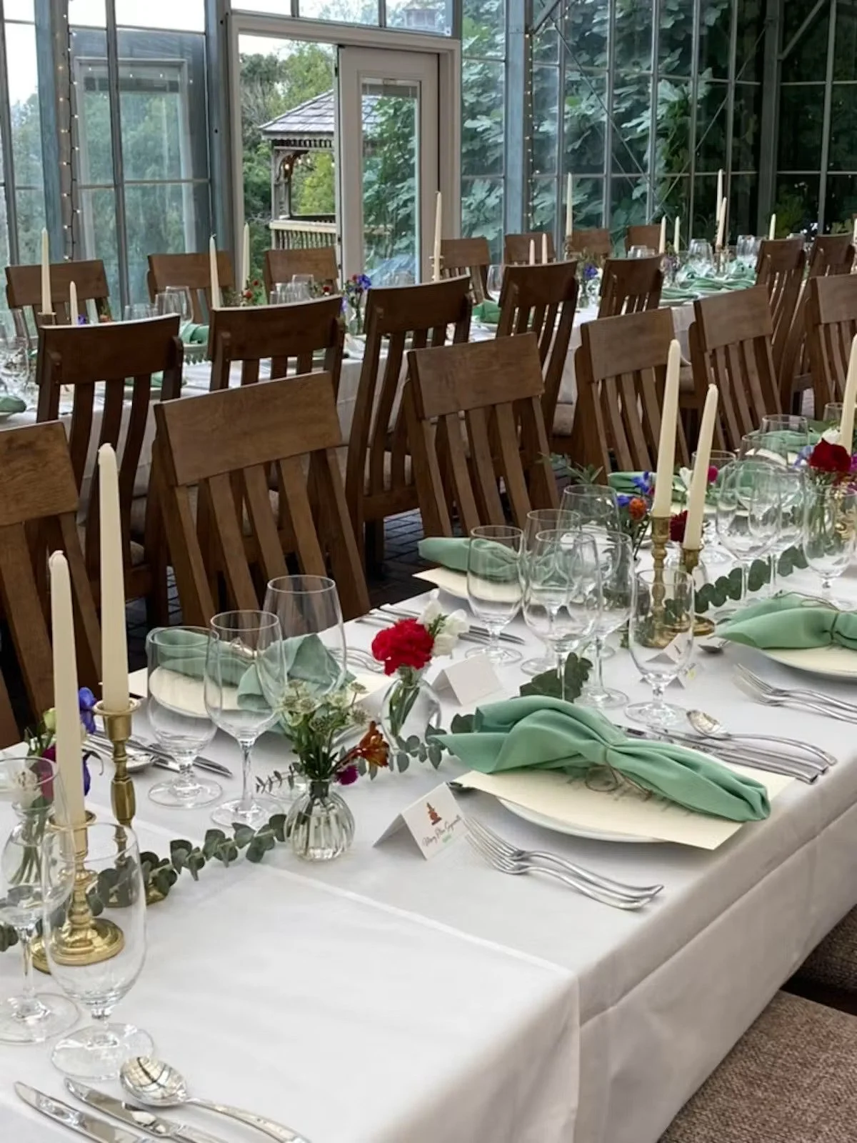 Indoor banquet table set with white tablecloth, green napkins, multiple wine glasses, and silverware, decorated with small flower arrangements, candles, and greenery, with wooden chairs around the table and a glass greenhouse structure with greenery 