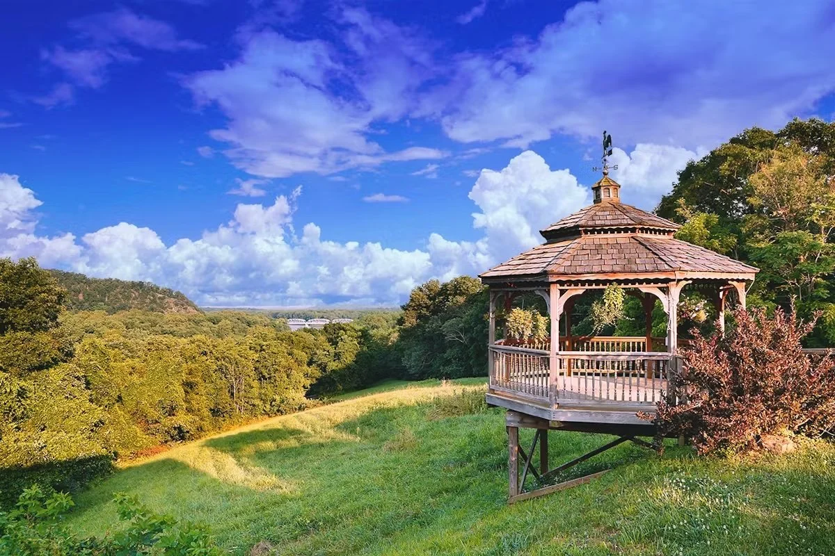 A wooden gazebo on a hillside overlooking a lush green river valley, under a bright blue sky with scattered clouds.