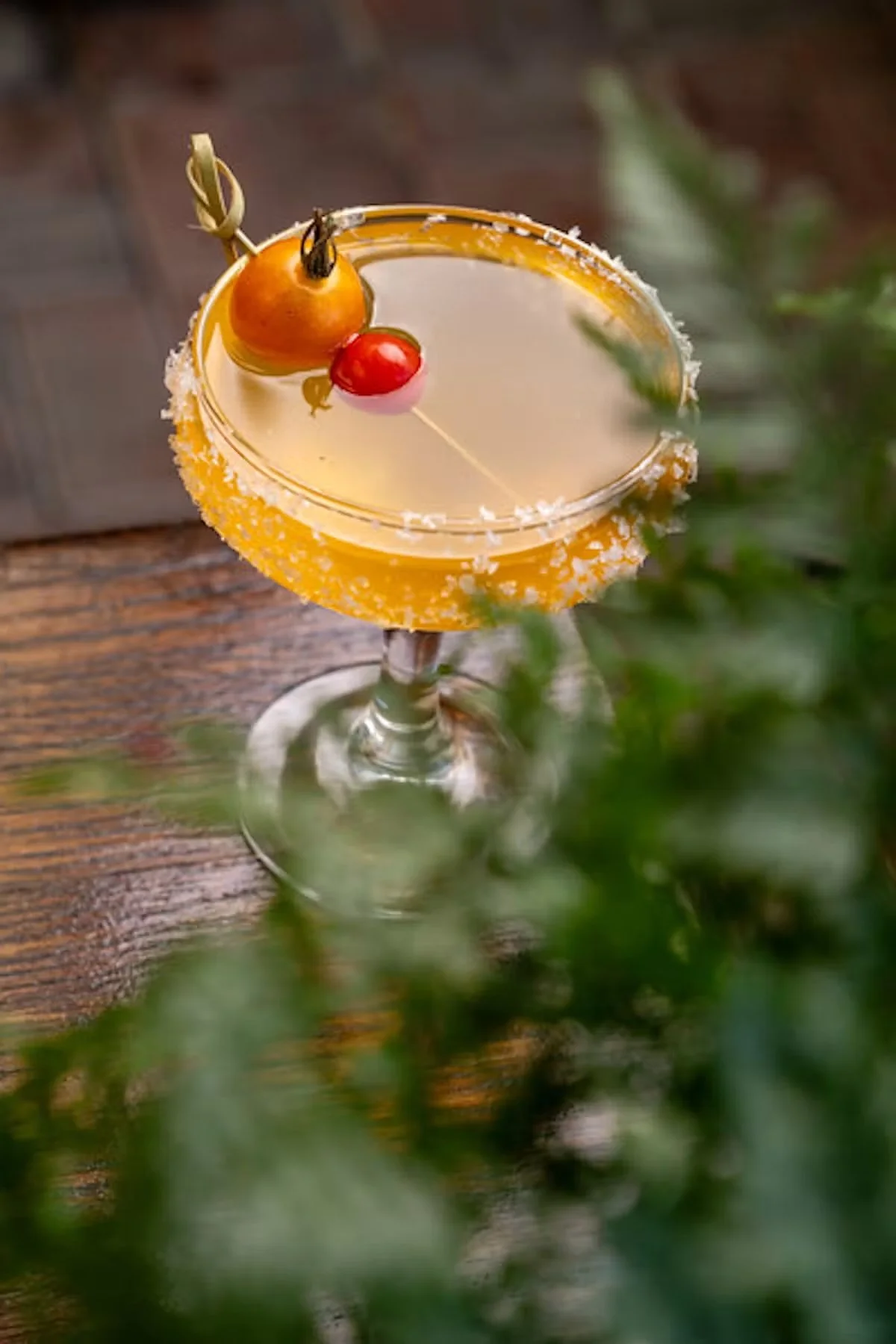 A cocktail in a stemmed glass with a salted rim, garnished with a cherry, a gooseberry, and a small lime wedge, placed on a wooden surface with out-of-focus greenery in the foreground.