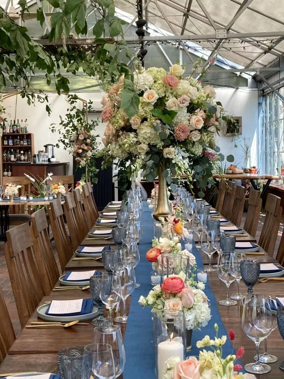 A long dining table decorated with a large floral centerpiece, set for a formal event in a rustic venue with a transparent ceiling. The table features multiple glasses, plates, napkins, and candles, with wooden chairs arranged on either side.