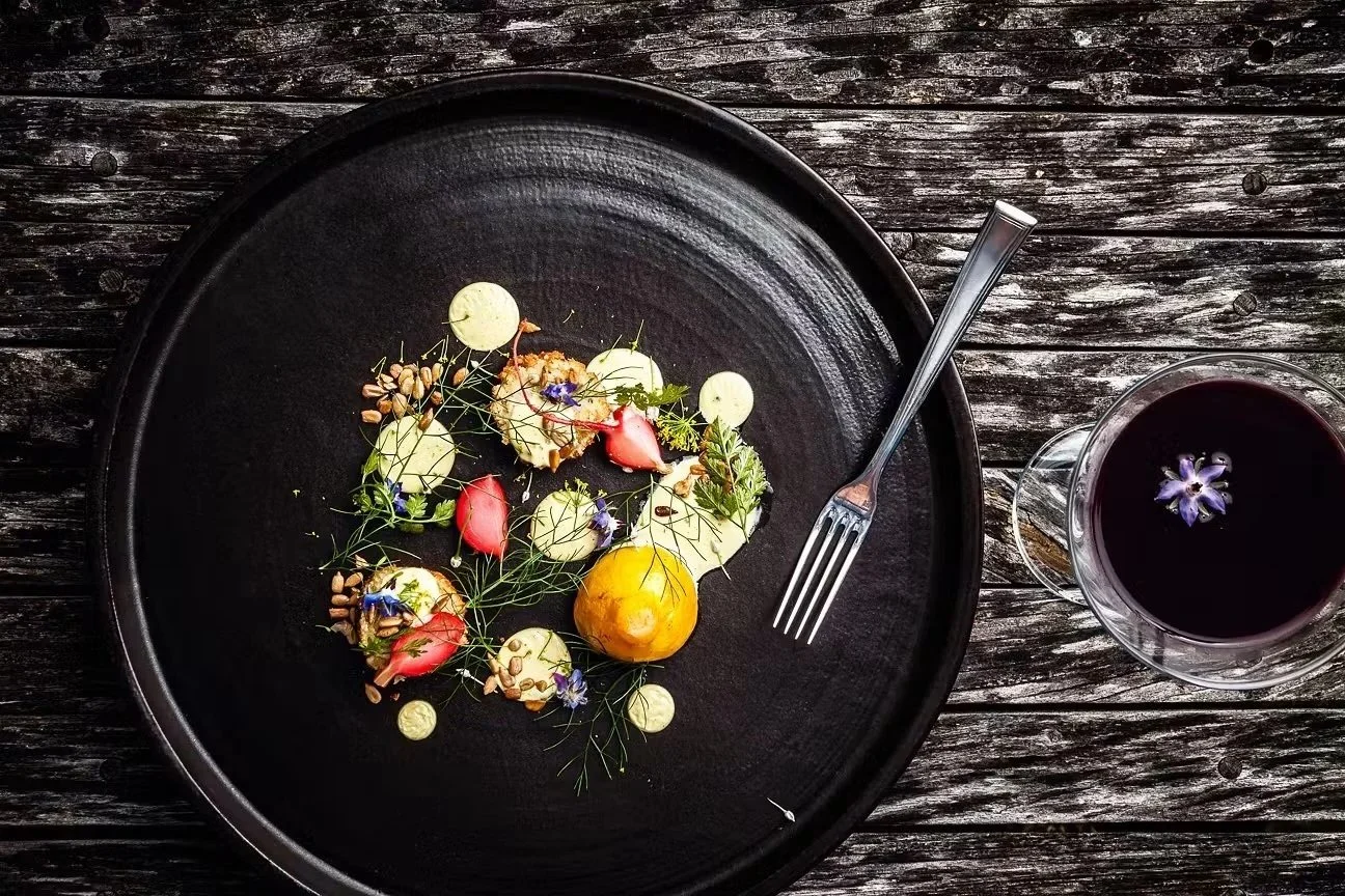 An overhead view of a black plate with artfully arranged small edible garnishes and a drizzle of white sauce. Next to the plate is a fork, and a glass of dark purple beverage with a purple flower in it is on a rustic wooden table.