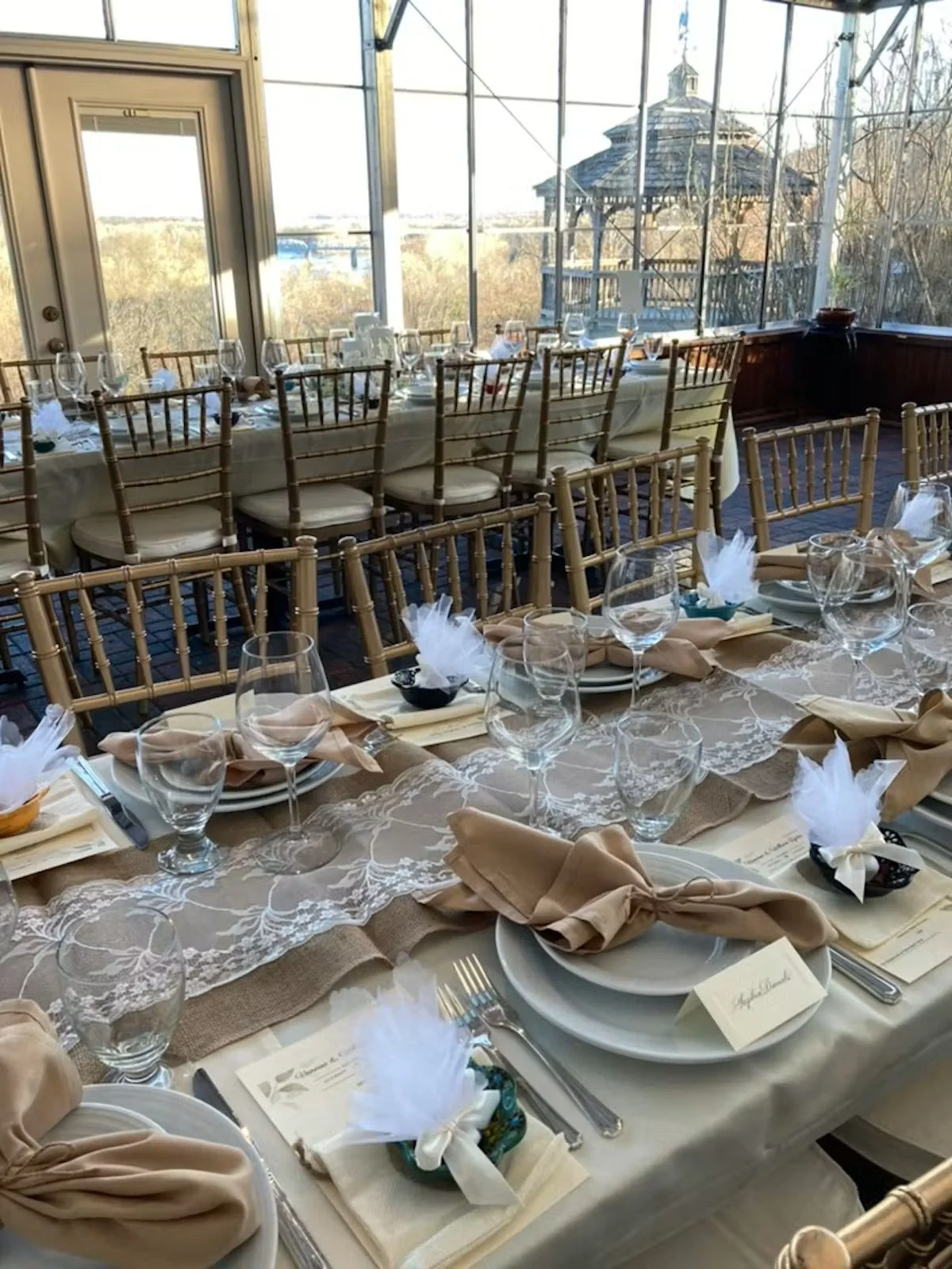 Elegant indoor dining setup with a long table, beige napkins, wine glasses, and place settings, with large windows overlooking an outdoor pavilion and scenic landscape.