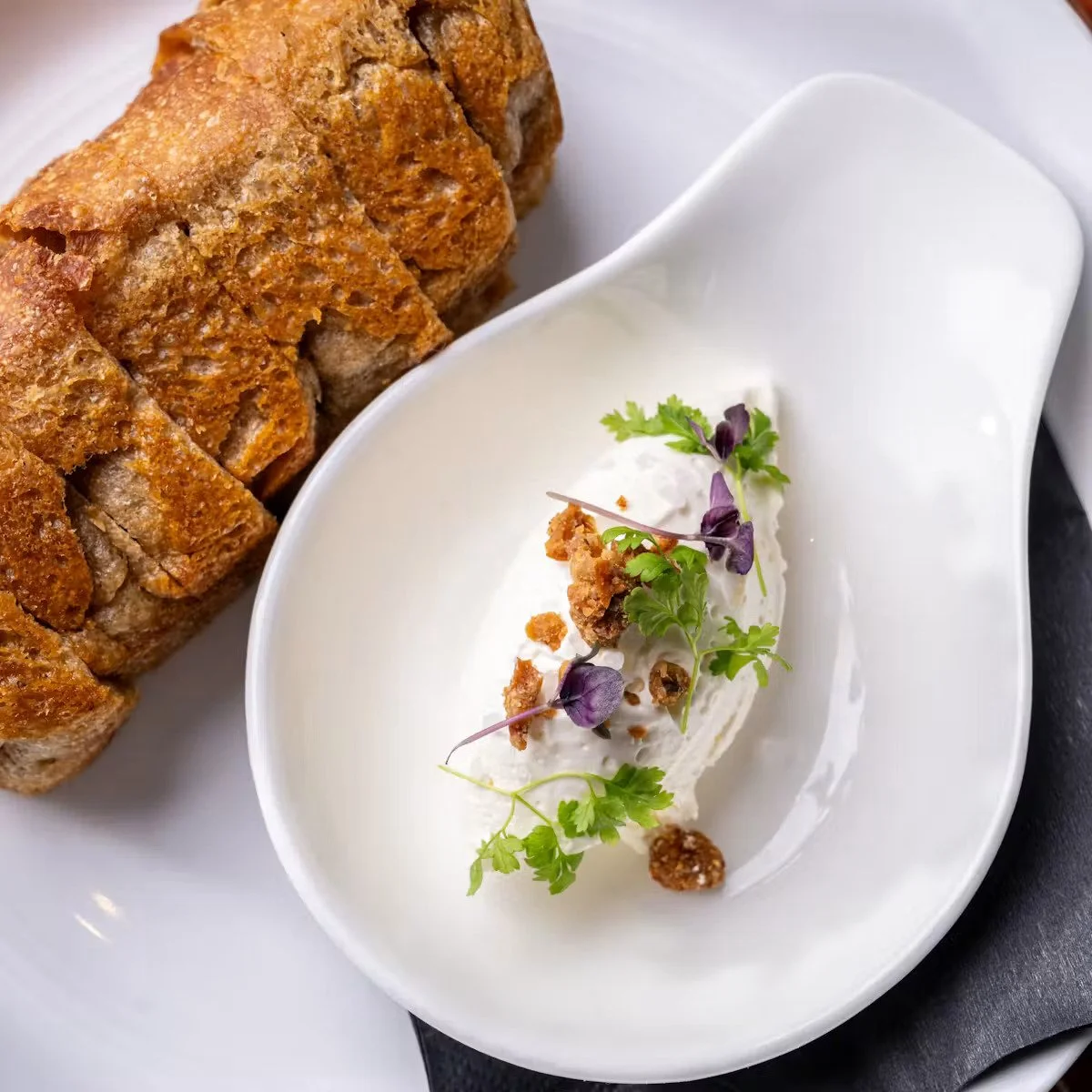 Close-up of a sliced loaf of bread next to a small white dish of creamy dip garnished with microgreens and crumbles.