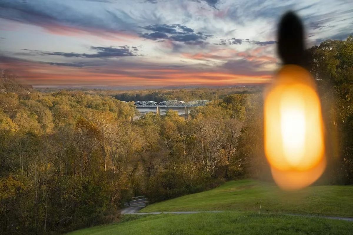 A scenic view of a sunset over a forest with a bridge in the distance. A blurred yellow-orange lantern is in the foreground.