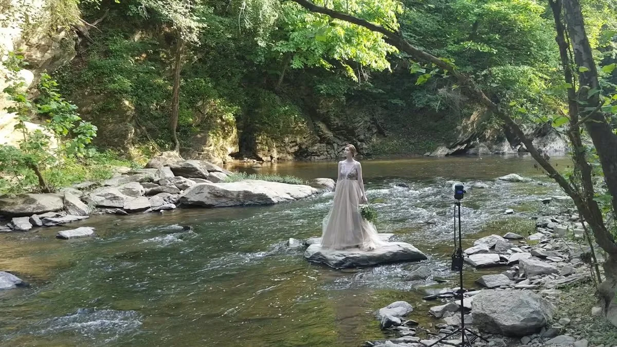 A woman in a white dress standing on a rock in the middle of a river, surrounded by green trees and rocky banks.