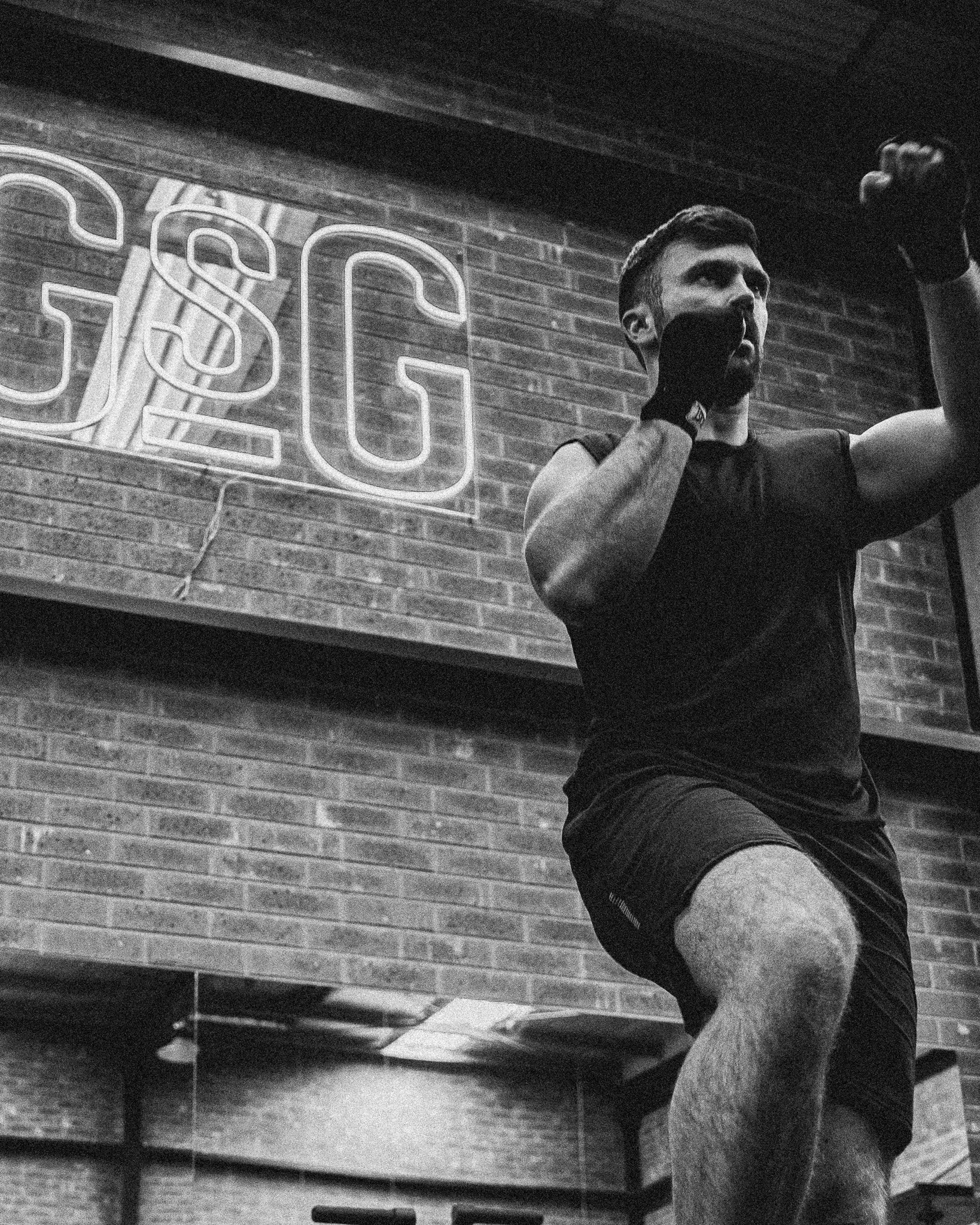 A man in athletic attire, wearing gloves, is in a gym performing a boxing move or shadow boxing in front of a mirror, with a large illuminated sign that reads 'ESG' on a brick wall behind him.