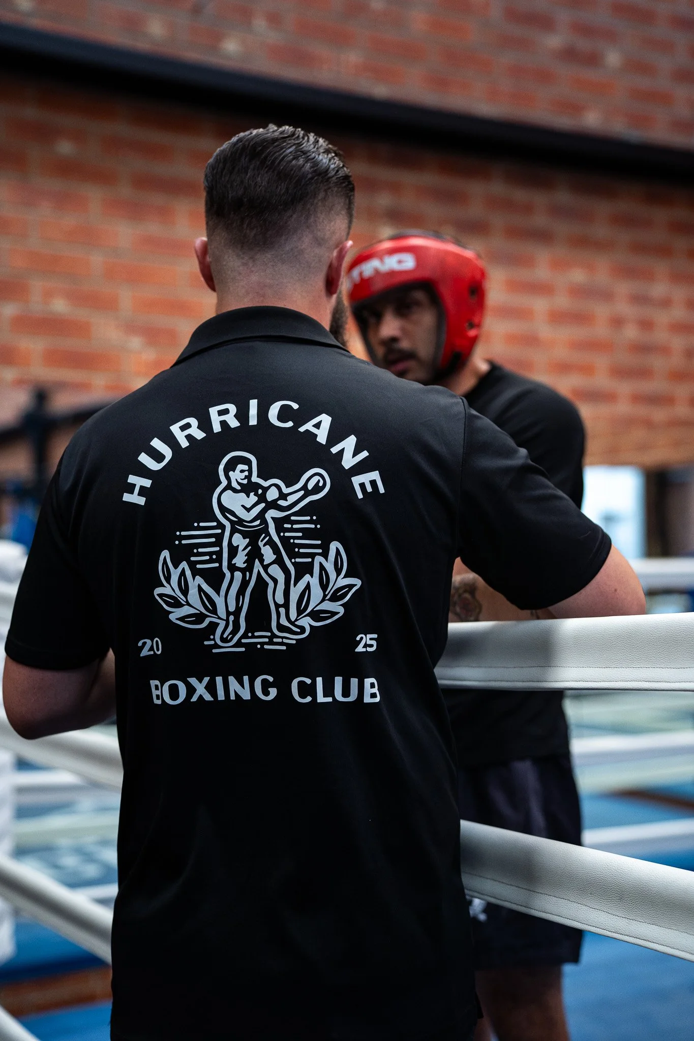 A man in a black t-shirt with 'HURRICANE BOXING CLUB' and a boxing graphic on the back, talking to a boxer wearing a red headgear inside a boxing gym.
