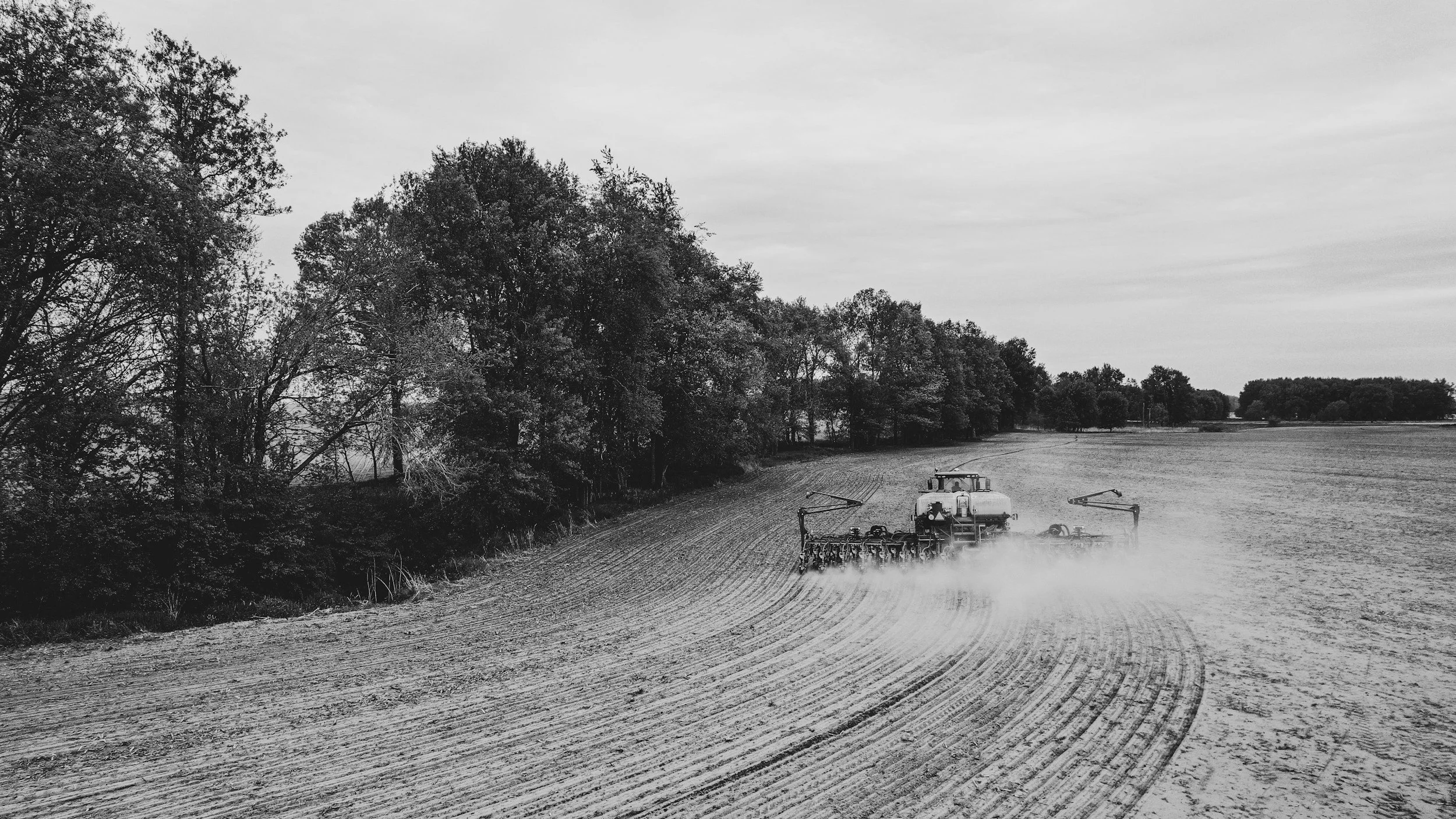 A field being tilled with a tractor that is creating dust, near a line of trees in the distance, in a rural setting on an overcast day. Drone Detection Ireland